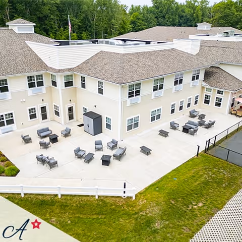 Aerial view of the outdoor patio area of a two-story assisted living facility building with multiple seating arrangements including chairs and tables on a concrete surface, surrounded by green grass and trees in the background.