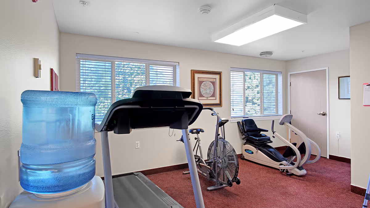 A small exercise room with a treadmill, stationary bike, and a recumbent exercise bike. The room has beige walls, red carpet, two windows with blinds, a water cooler, and framed artwork on the walls.