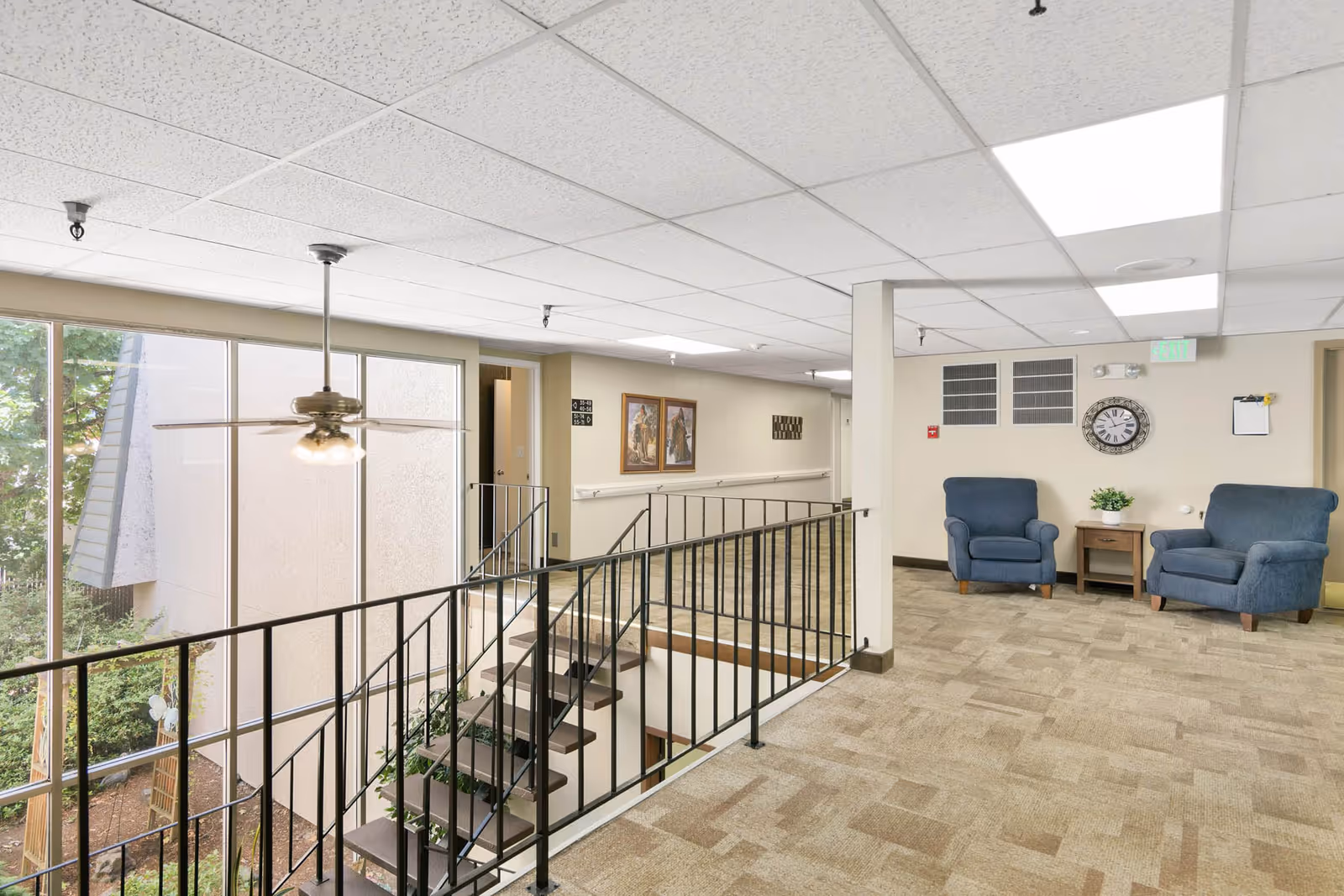 Interior view of a senior living facility hallway with a staircase leading downstairs, two blue armchairs with a small wooden table between them, a wall clock, framed artwork, and large windows showing outdoor greenery.