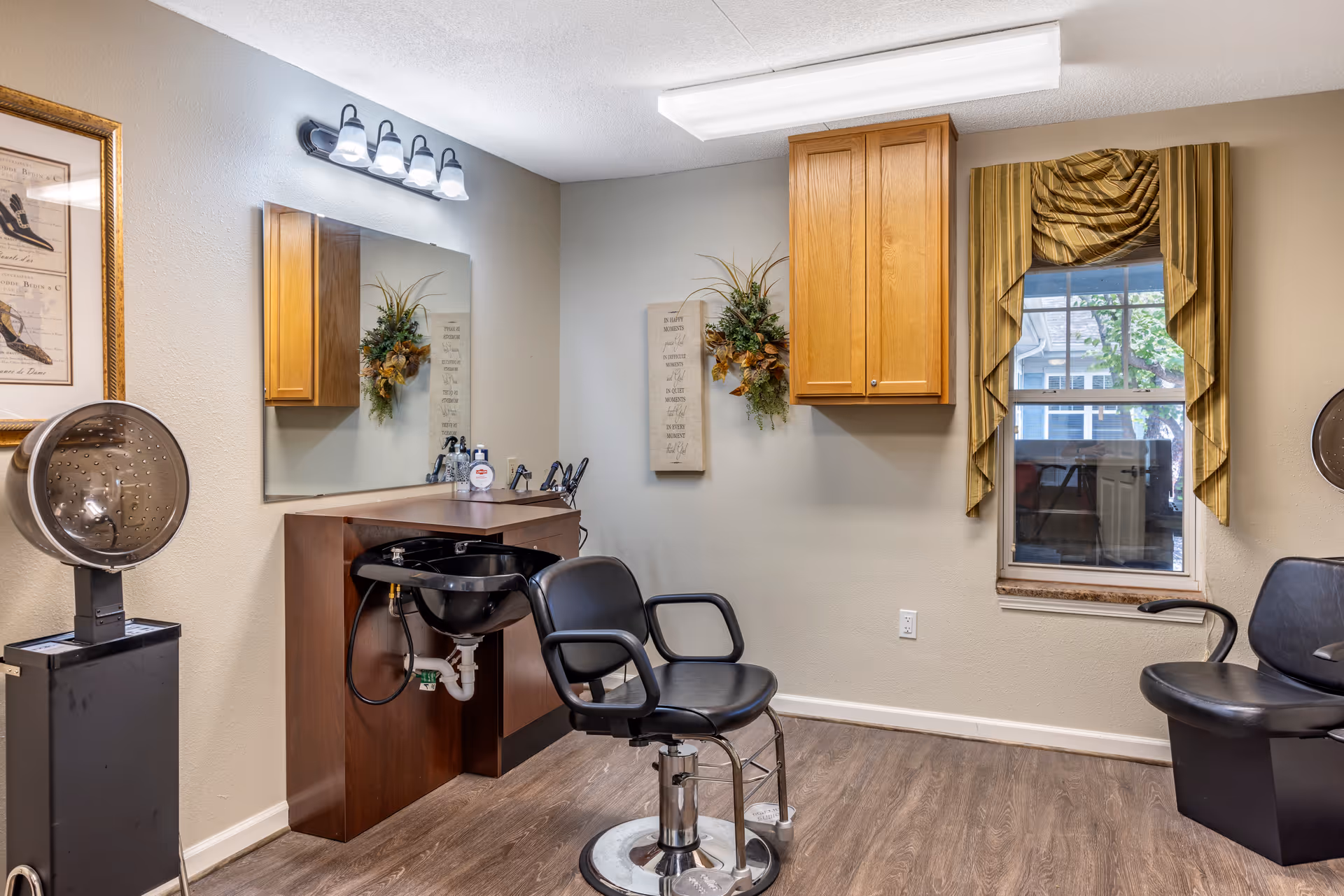 A small salon room with two styling chairs, a shampoo sink and counter, mirror and wooden cabinets beside a window with draped curtains.