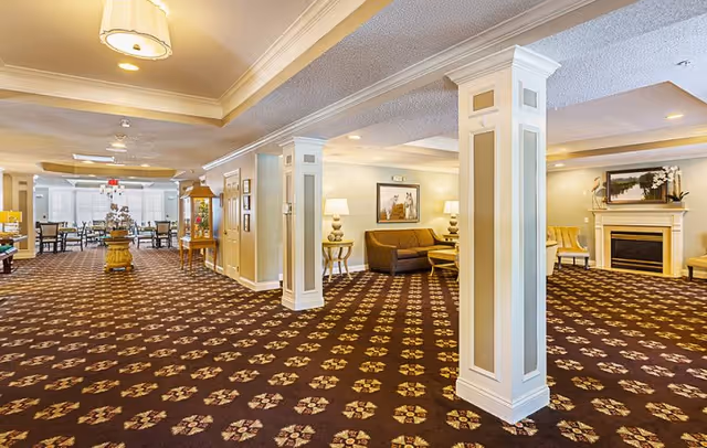 Spacious interior common area with patterned carpet, white columns, and soft lighting. The room features seating areas with sofas and chairs, a fireplace with artwork above it, and a dining area with tables and chairs in the background.