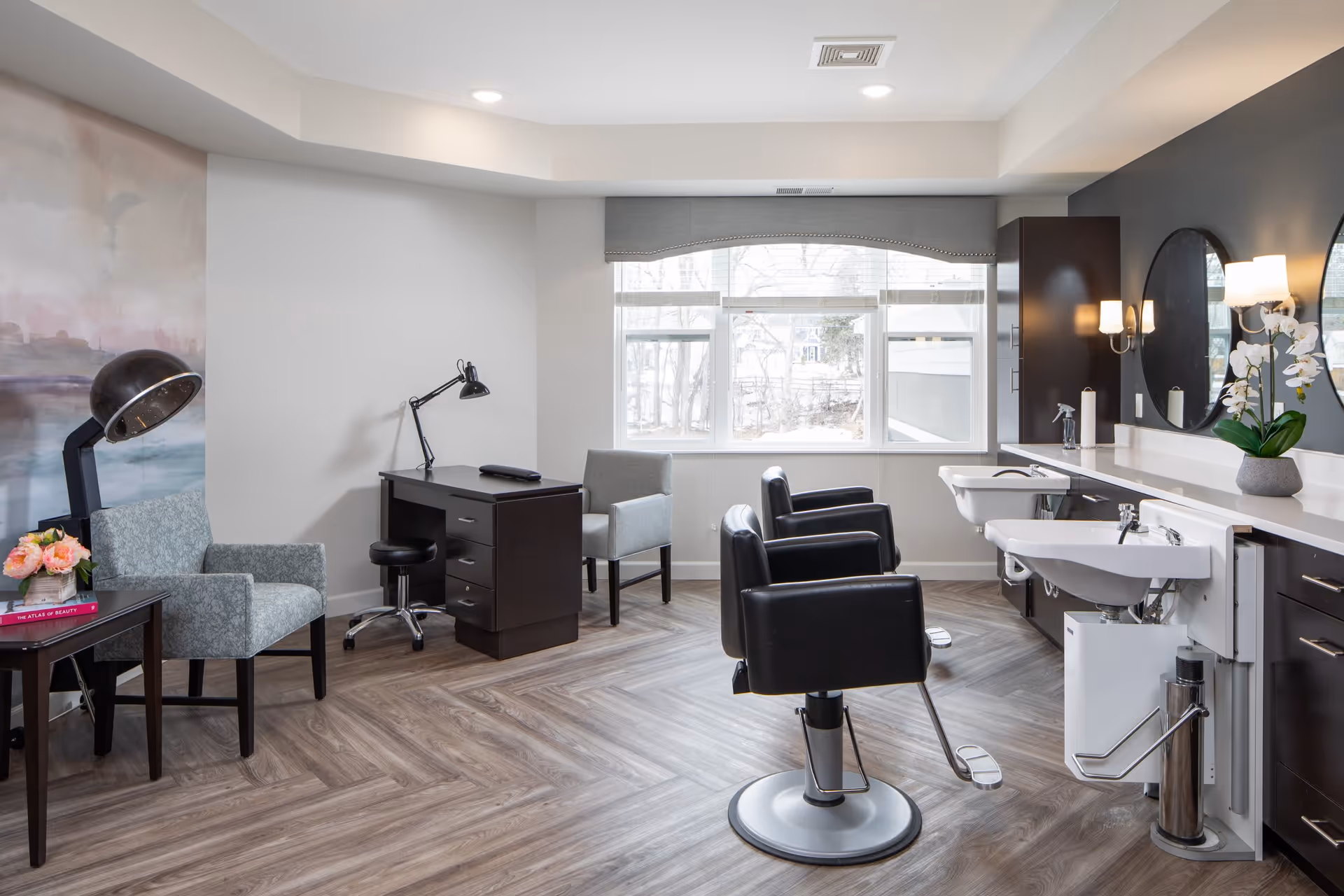 A bright and modern salon room with two black salon chairs in front of two white sinks and large round mirrors mounted on a dark accent wall. The room has wood-patterned flooring, a window with a gray valance letting in natural light, a small desk with a lamp, and two upholstered chairs. A vintage hair dryer and a small table with flowers and a book are also visible.
