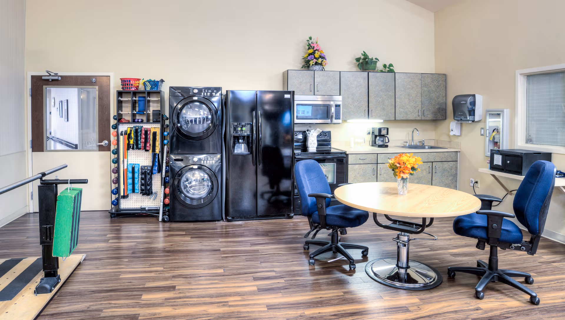 A multi-purpose room with a kitchenette and laundry area. The room features a stacked washer and dryer, a black refrigerator, a microwave, an oven, and cabinets. There is a round wooden table with a vase of flowers and two blue office chairs on wheels. The floor is wood laminate, and there is a rack with exercise equipment next to the washer and dryer. A door with a window is visible on the left side, and a window with blinds is on the right.