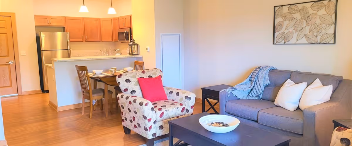 A cozy living room and kitchen area in an apartment. The living room has a gray sofa with white pillows and a blue throw blanket, a patterned armchair with a red pillow, a dark wooden coffee table with a white bowl, and a side table. The kitchen features wooden cabinets, a stainless steel refrigerator, a microwave, and a small dining table with two chairs set with dishes and cups. The floor is wooden, and there is a framed leaf artwork on the wall above the sofa.