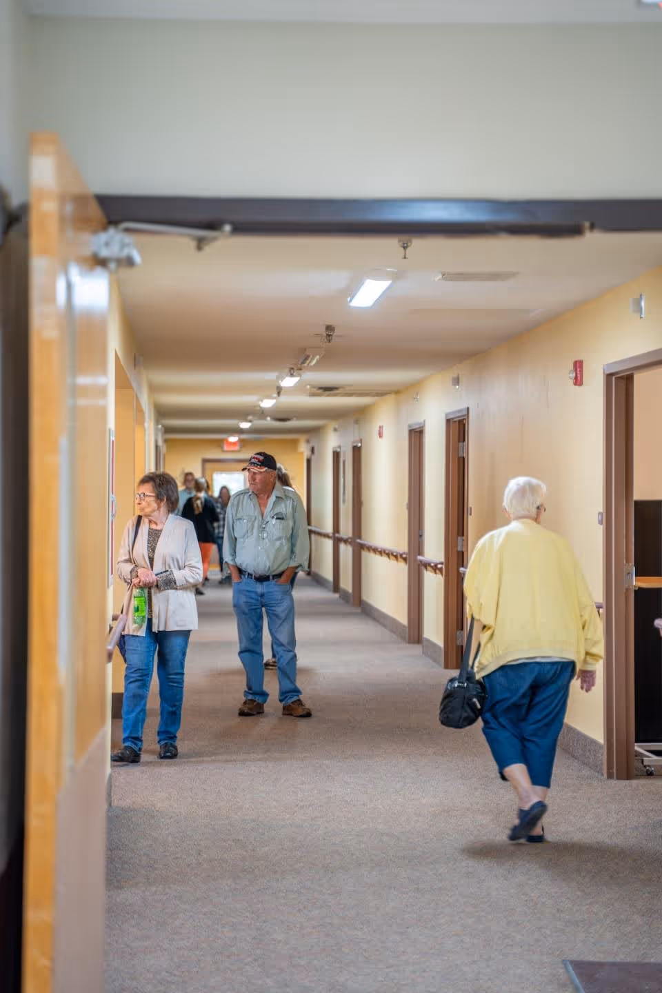 A long indoor hallway in Eagles Rest Assisted Living with beige walls and carpeted floor. Several elderly people are walking or standing in the hallway near the doors to rooms. The hallway is well-lit with ceiling lights and has handrails along the walls.