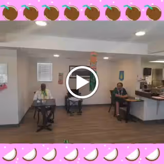Interior common room with three people seated at tables playing electronic keyboards in a bright community area with wood floors and a pink decorative border.