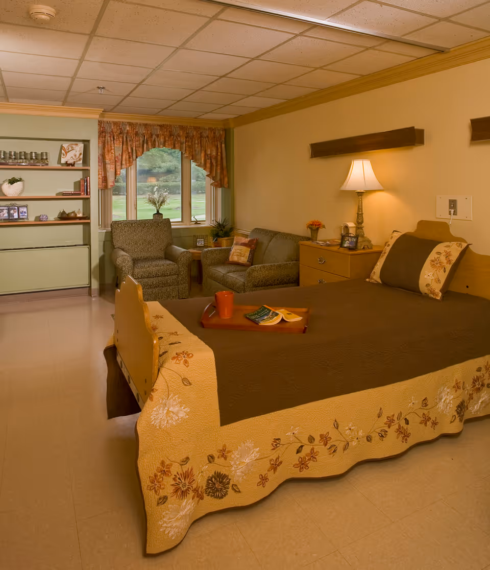 A cozy senior living room with a bed covered in a brown and floral quilt, a wooden nightstand with a lamp and small decorations, a comfortable armchair and loveseat near a window with floral curtains, and built-in shelves with decorative items and framed photos.