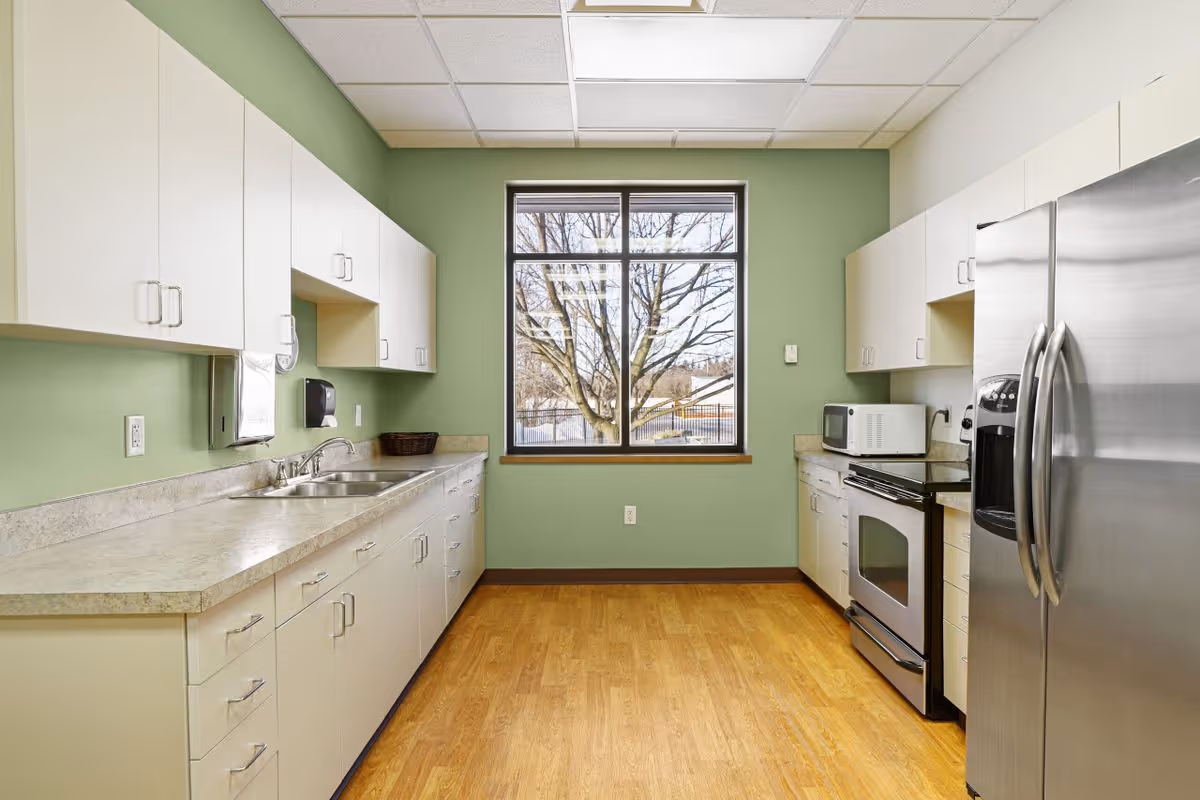 Bright institutional kitchen with white cabinets, a double sink along the left countertop, a stainless steel refrigerator and stove on the right, and a central window on a green wall.