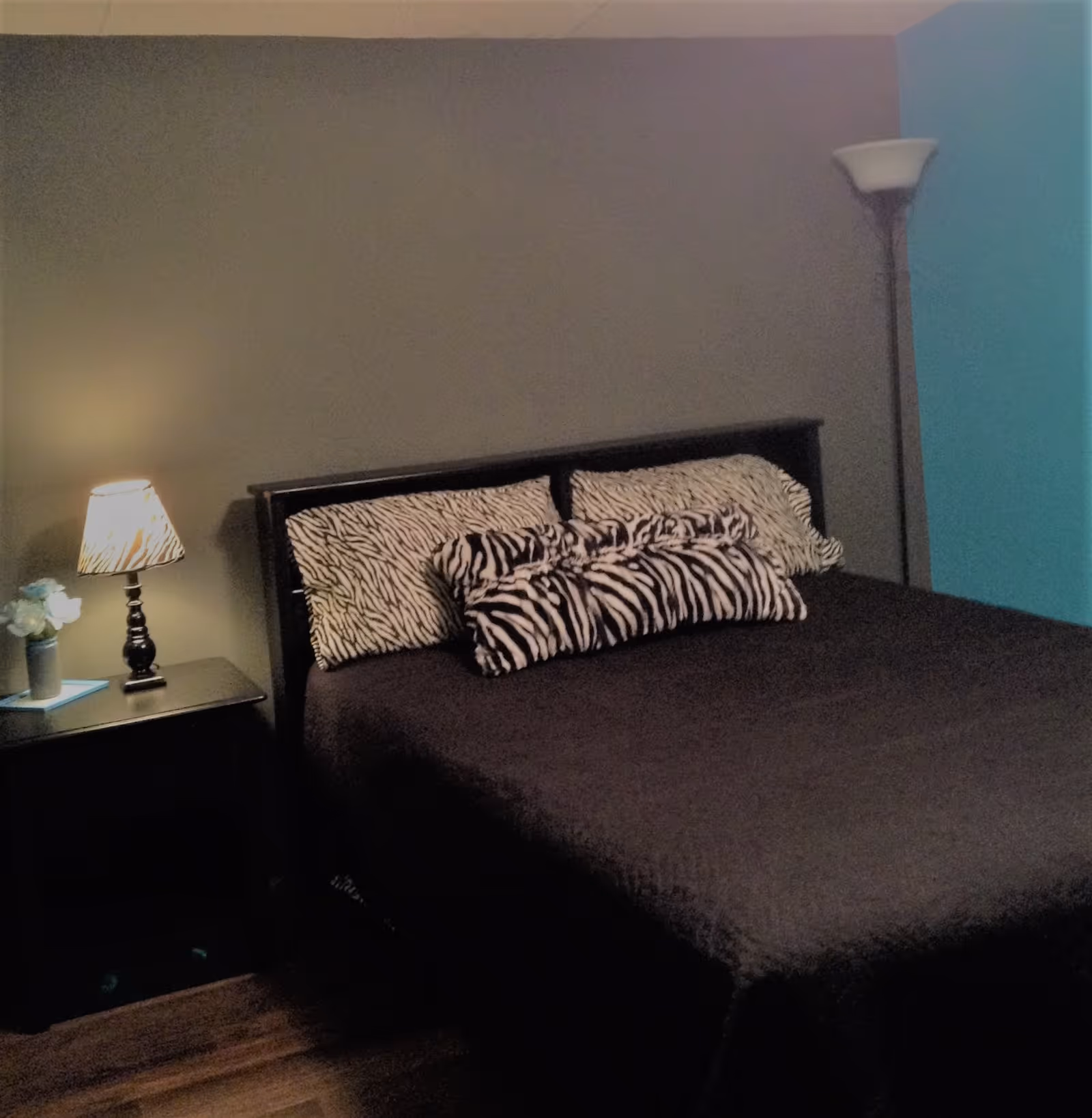A bedroom with a dark bedspread on a bed, decorated with zebra-patterned pillows. There is a nightstand with a lamp and a small vase with flowers on the left side of the bed. A tall floor lamp stands in the corner against a blue accent wall.
