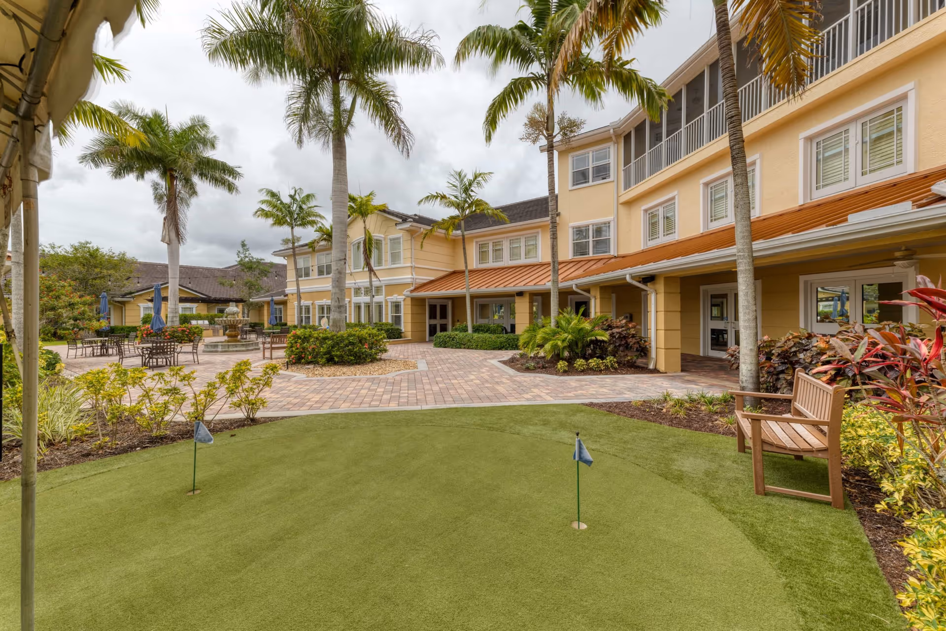 Outdoor courtyard area of a senior living facility with palm trees, a small putting green with two flags, benches, patio tables with umbrellas, and a multi-story yellow building in the background under a cloudy sky.
