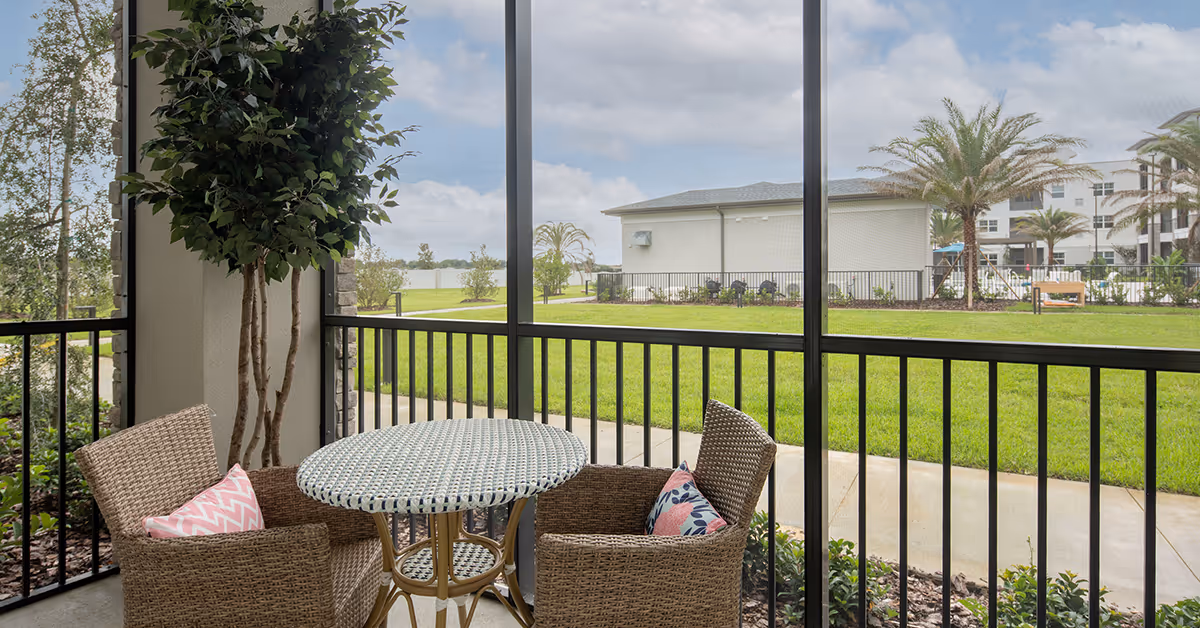 A screened-in patio area with a round wicker table and two wicker chairs, each with a decorative pillow. The patio overlooks a green lawn with palm trees and a building in the background under a partly cloudy sky.