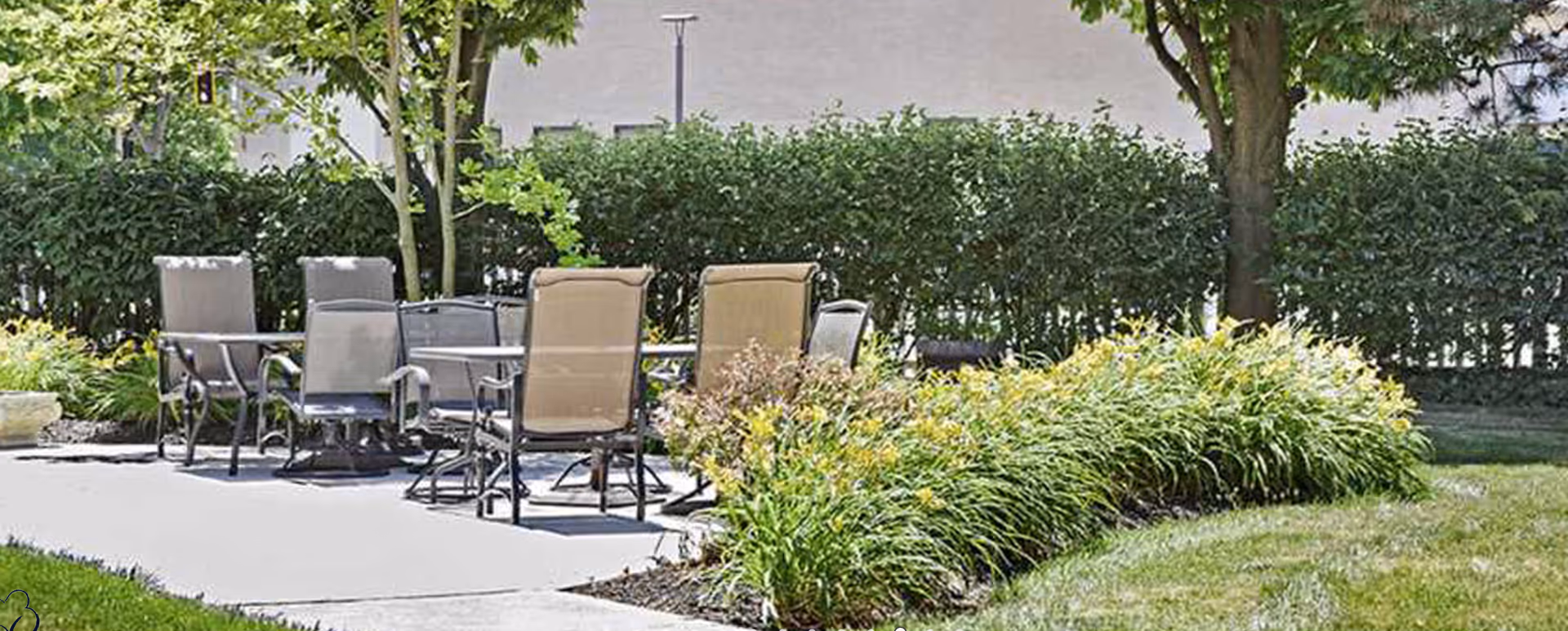 Outdoor patio area with several metal and mesh chairs arranged around two tables, surrounded by green bushes, trees, and landscaped plants under daylight.