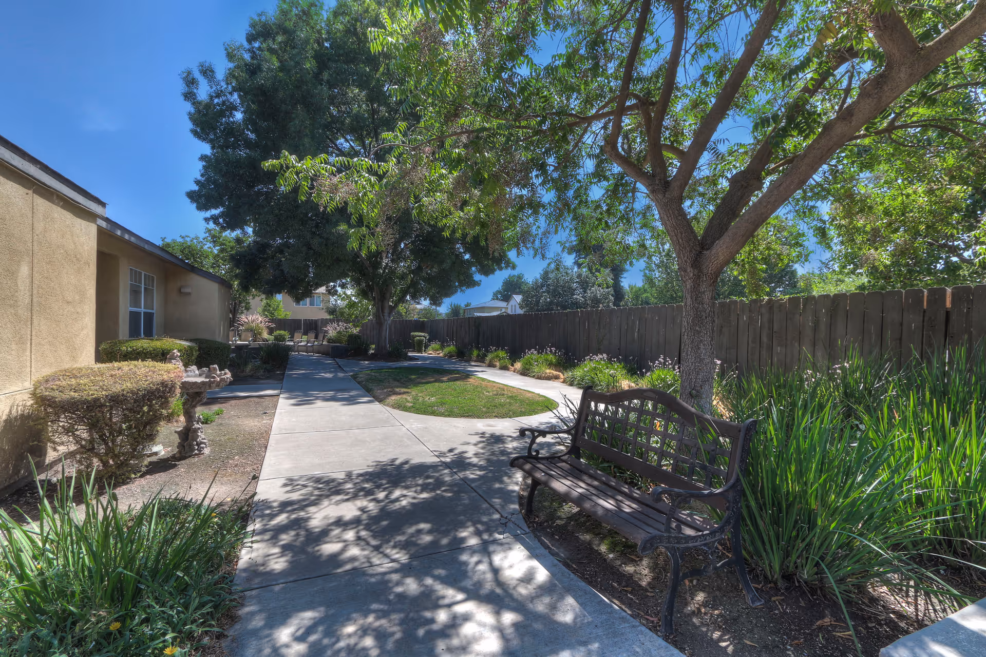 Outdoor garden area at The Gardens of Modesto featuring a paved walkway, a wooden bench under a tree, green shrubs, and a wooden fence under a clear blue sky.
