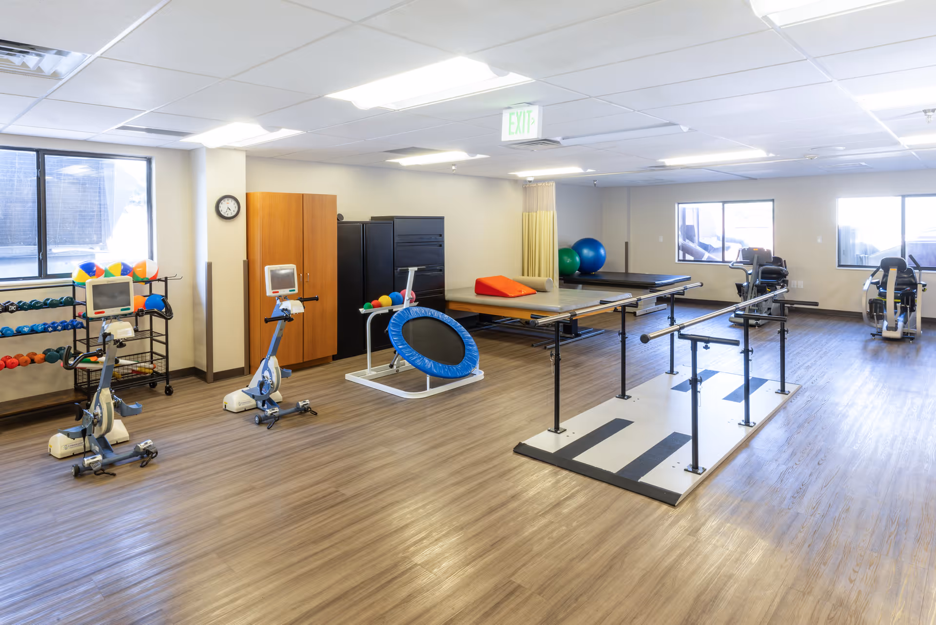 Bright physical therapy and exercise room with parallel bars, exercise bikes, mats, weights and therapy equipment.