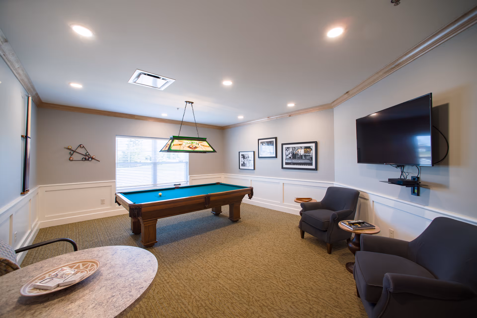 A well-lit game room featuring a pool table with a green felt surface, two gray armchairs, a wall-mounted flat-screen TV, and framed black-and-white photographs on the walls. The room has beige carpeting, white wainscoting, and a window with blinds letting in natural light.