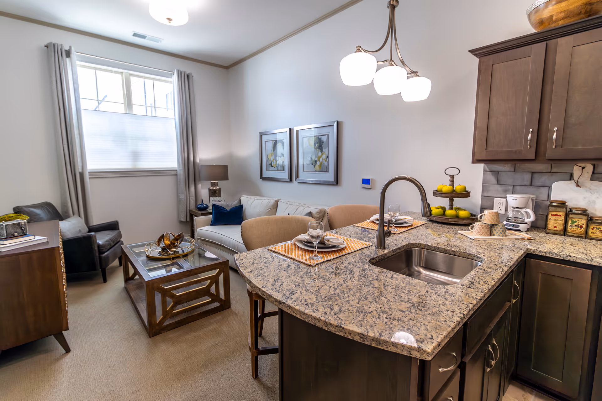 A cozy living room and kitchen area in a senior living facility. The living room features a white sofa with blue and beige pillows, a dark leather armchair, a wooden coffee table with decorative items, and two framed floral pictures on the wall. The kitchen has a granite countertop with a built-in sink, two place settings with plates and glasses, a tiered fruit stand with green apples, a coffee maker, and several jars. The room is well-lit with natural light from a window with gray curtains and a ceiling light fixture above the kitchen counter.