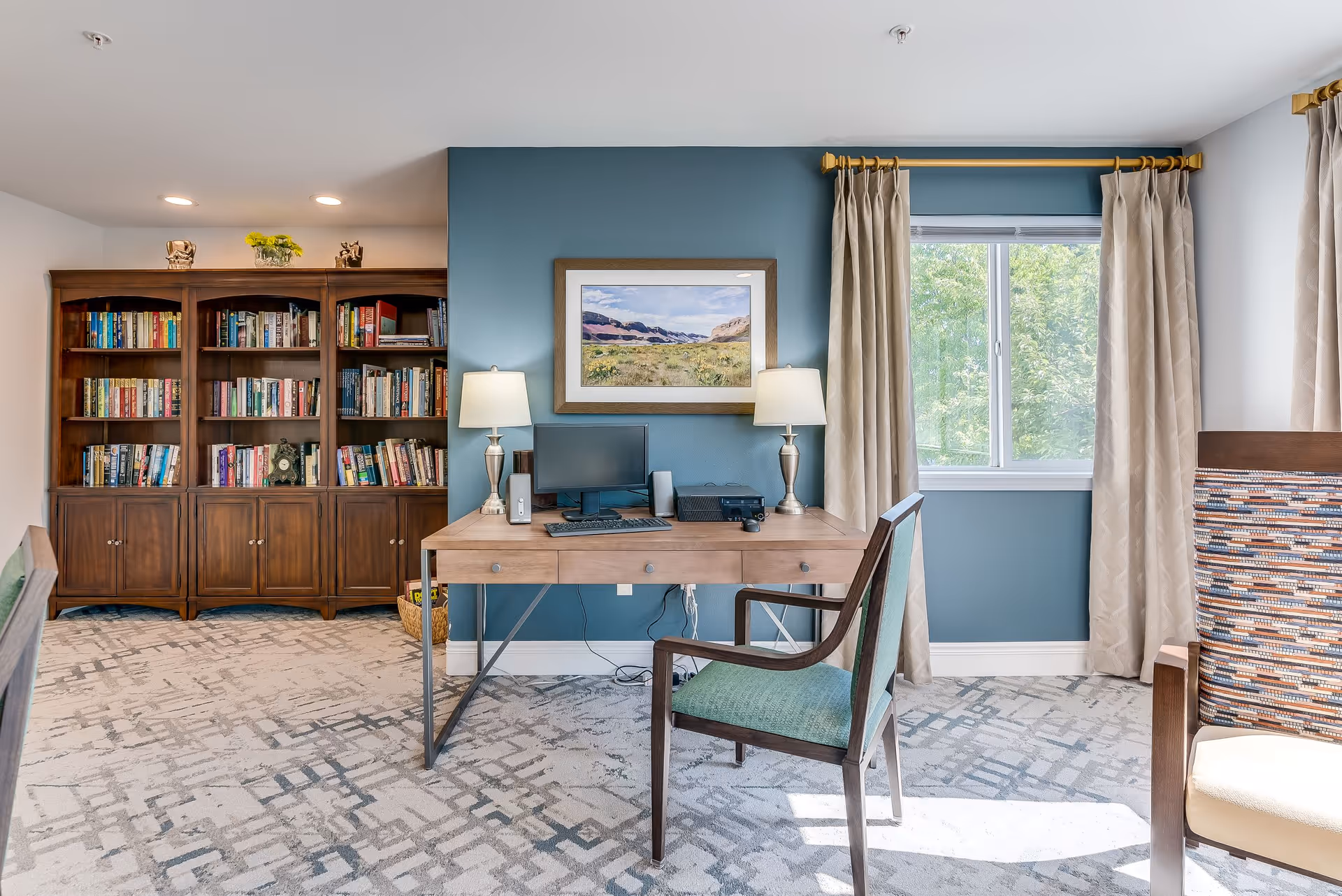 A bright and cozy interior room featuring a wooden desk with a computer, two table lamps, and a chair with green upholstery. Behind the desk is a blue accent wall with a framed landscape painting. To the left, there is a large wooden bookshelf filled with books. A window with beige curtains allows natural light to enter the room, and part of another chair with a patterned backrest is visible on the right side.