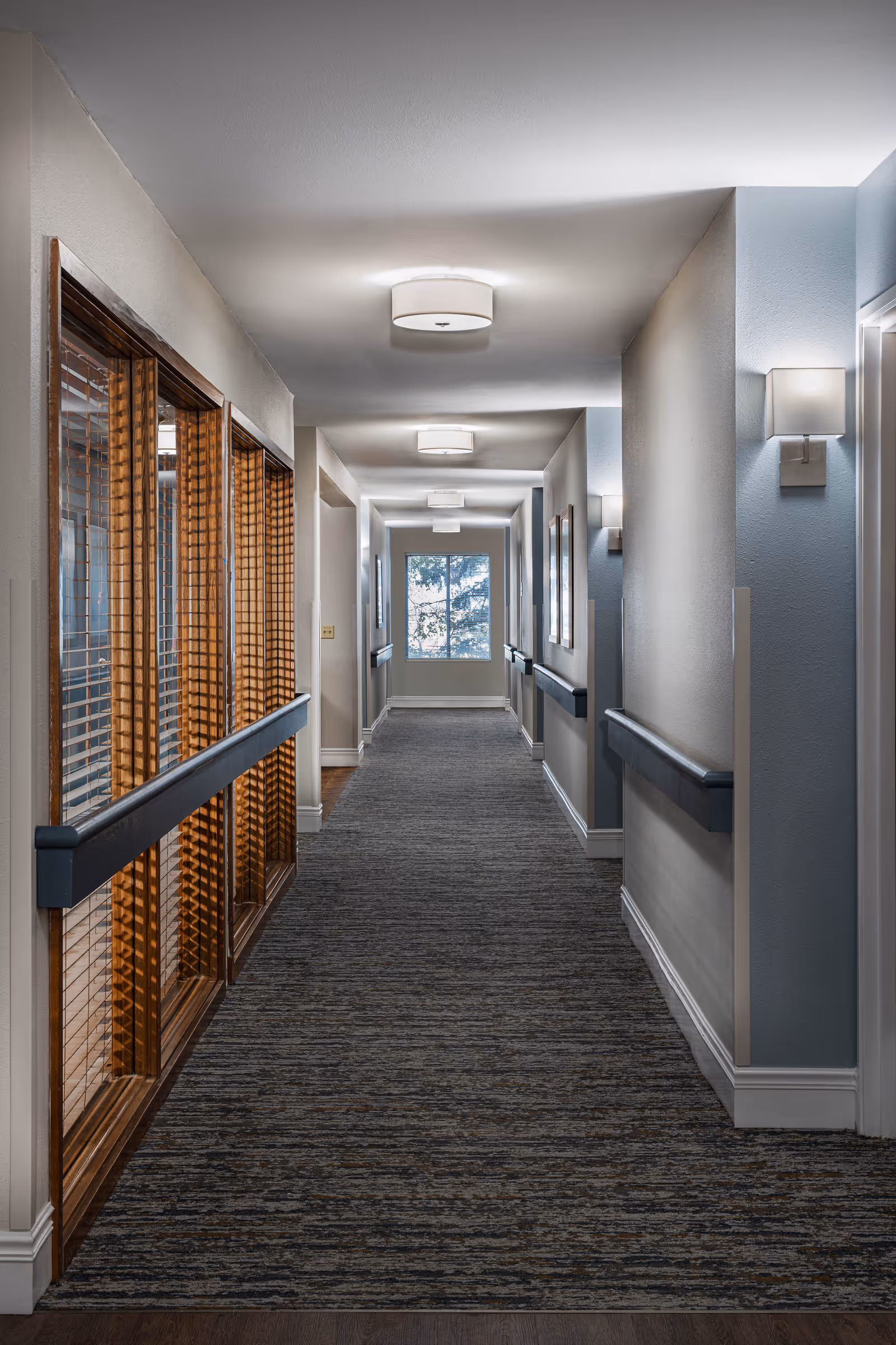 A well-lit indoor hallway in a senior living facility with carpeted floor, handrails on both sides, wall-mounted lights, and a window at the far end letting in natural light. Wooden framed windows with blinds are on the left side.
