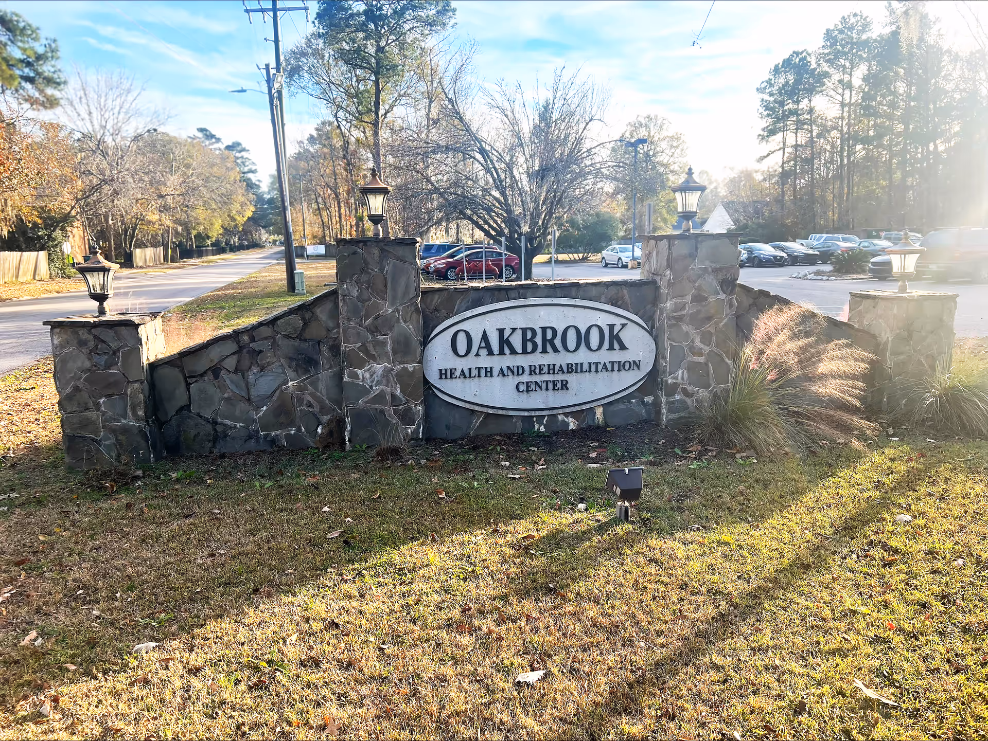 Stone sign for Oakbrook Health and Rehabilitation Center with four stone pillars topped with lanterns, situated on a grassy area near a road with trees and parked cars in the background.