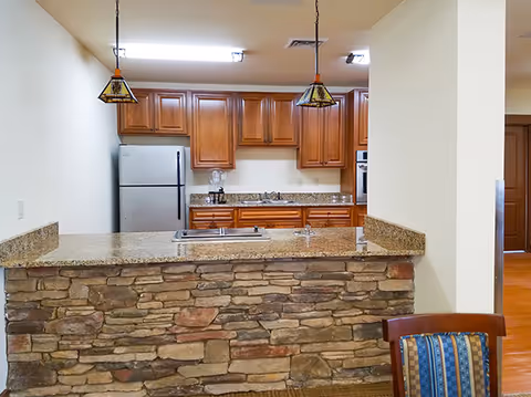 Interior view of a kitchen area in a senior living facility featuring wooden cabinets, a stainless steel refrigerator, a built-in oven, a granite countertop with a stone facade, two pendant lights hanging from the ceiling, and a chair with a blue patterned cushion partially visible in the foreground.