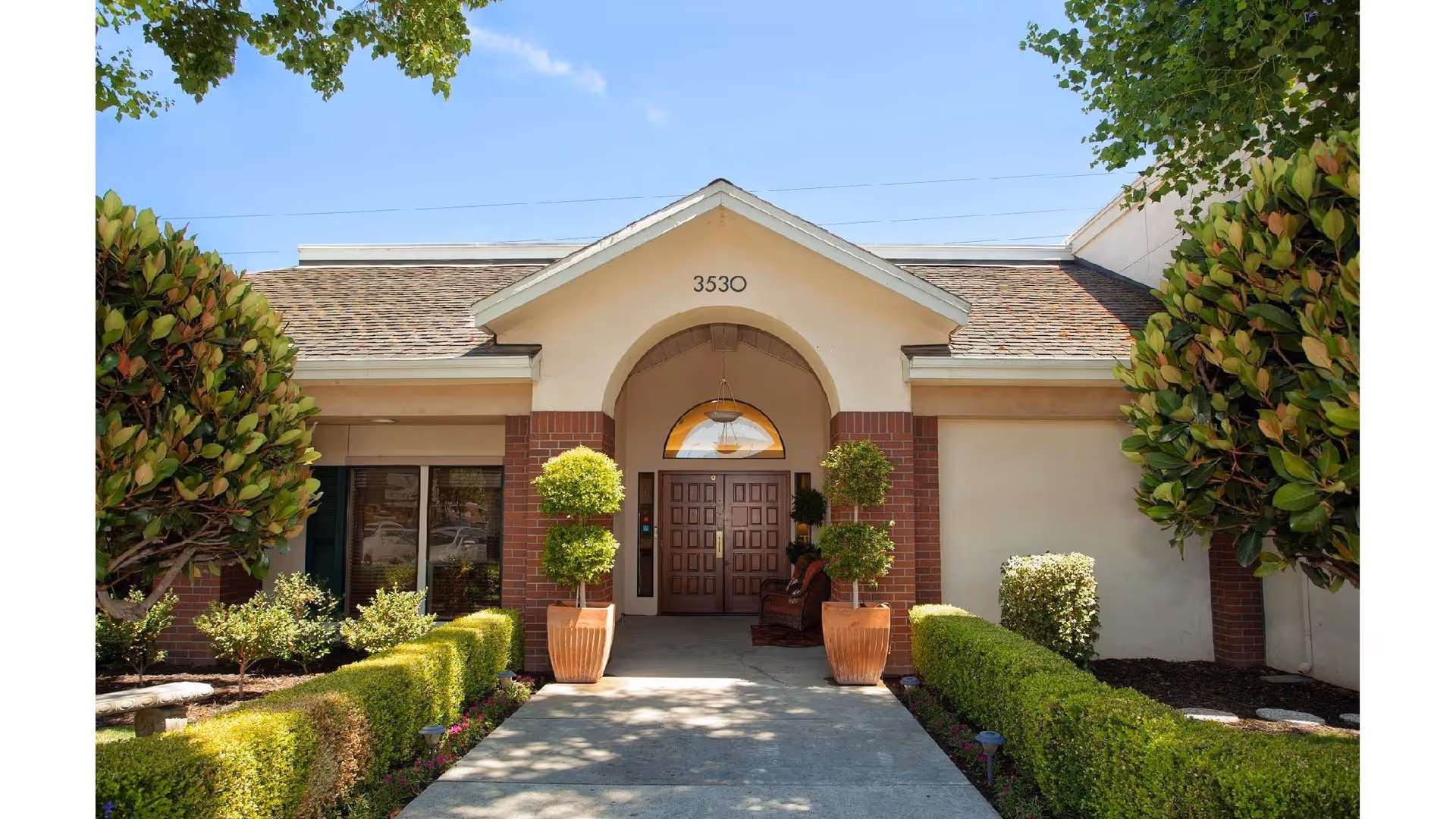 Front entrance of a single-story building with a peaked roof and the number 3530 above the doorway. The entrance is framed by two large potted plants with trimmed greenery, and there are manicured hedges and trees on either side of the concrete walkway leading to the door. The sky is clear and blue.