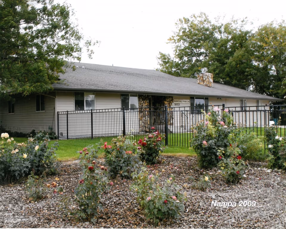 Single-story building with beige siding and a stone chimney, surrounded by a black metal fence and a garden with various rose bushes. Trees are visible in the background under an overcast sky.