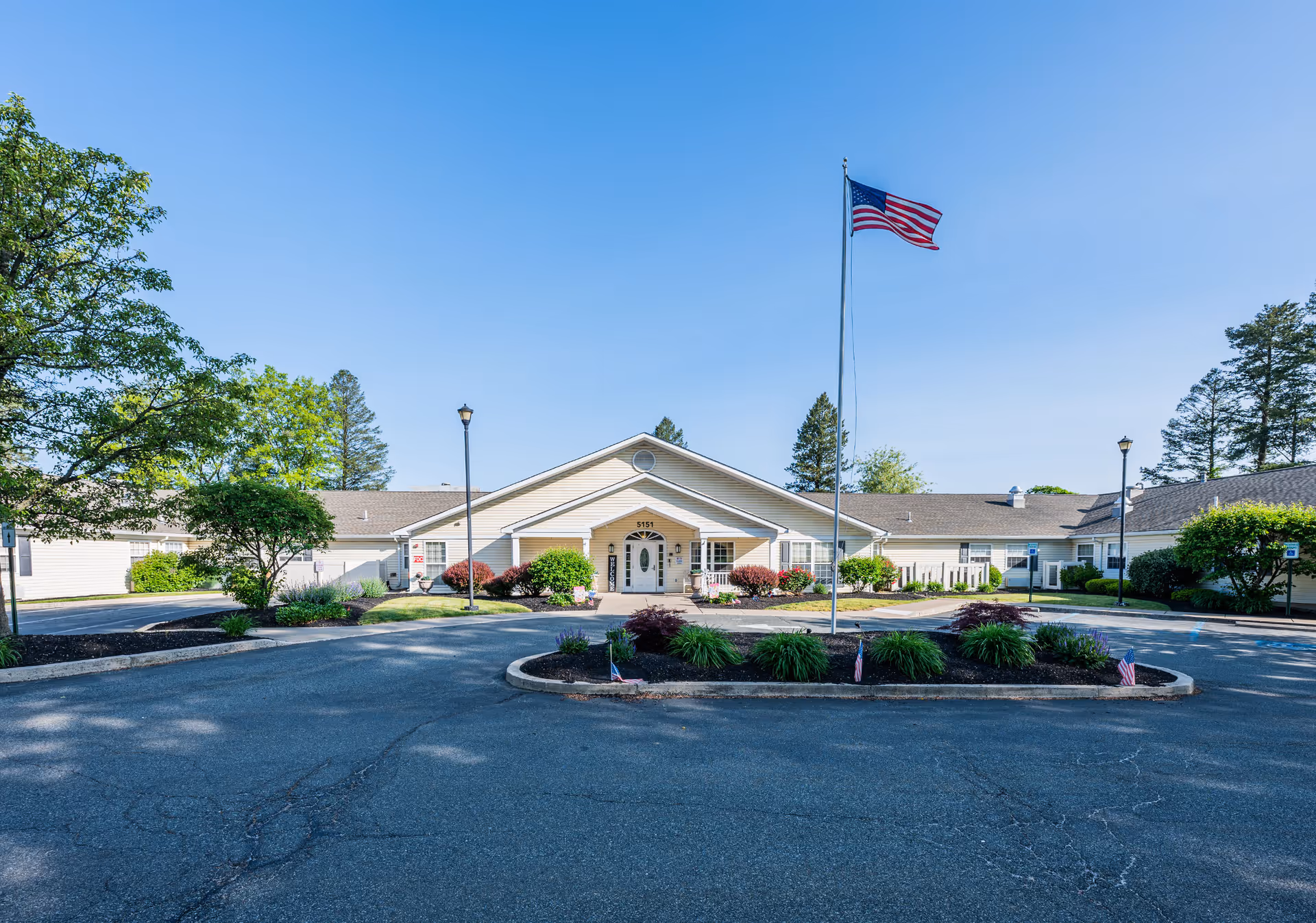 Front exterior view of Arden Courts - ProMedica Memory Care Community in Allentown, showing a single-story building with a peaked roof, an American flag on a flagpole in the center, landscaped greenery, and a circular driveway.