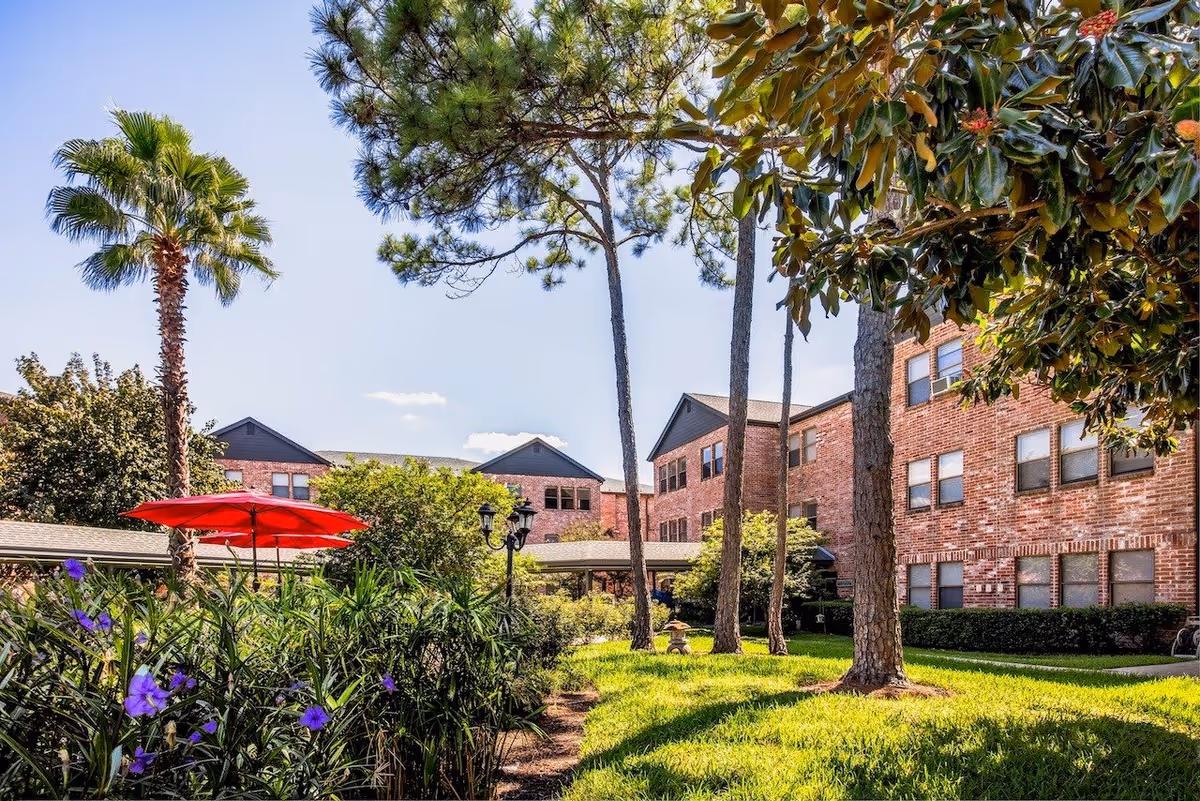 Outdoor garden area at Tarrytowne Estates featuring green grass, tall trees including palm trees, flowering plants, a red patio umbrella, and a brick building in the background under a clear blue sky.