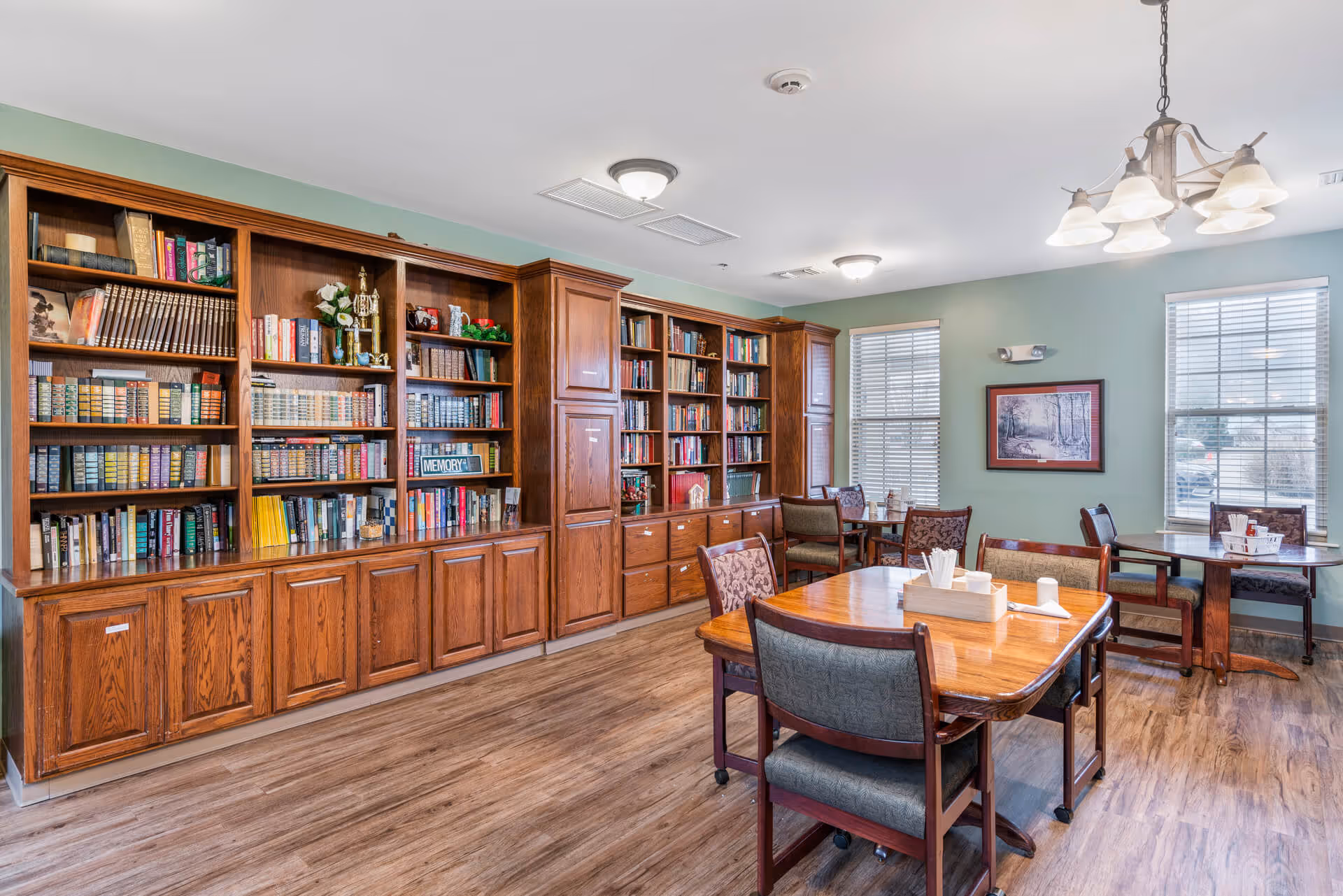 Bright common room with large wooden bookshelves along one wall, wooden tables and chairs, and windows letting in natural light.