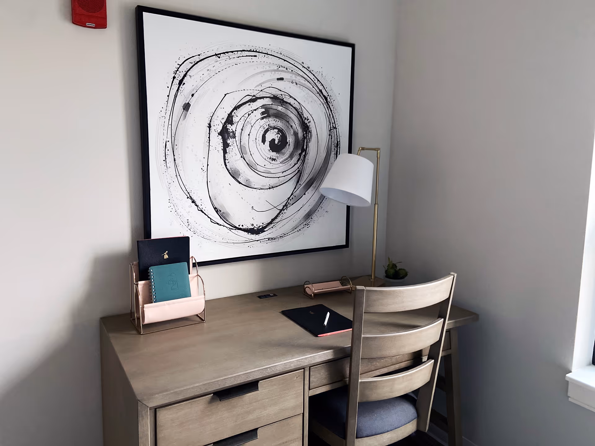 A wooden desk with a matching wooden chair in front of a wall. On the desk are a desk lamp, a small plant, a black notebook with a pen, a pink organizer holding two notebooks, and a large black and white abstract circular painting hanging on the wall above the desk.