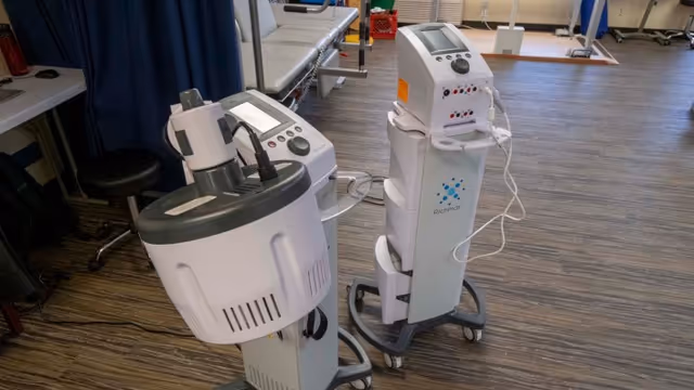 A medical treatment room with two pieces of medical equipment on wheels in the foreground. The room has wood-patterned flooring, a treatment table with a gray cushion, a black stool, and a blue privacy curtain. Various medical supplies and equipment are visible in the background.