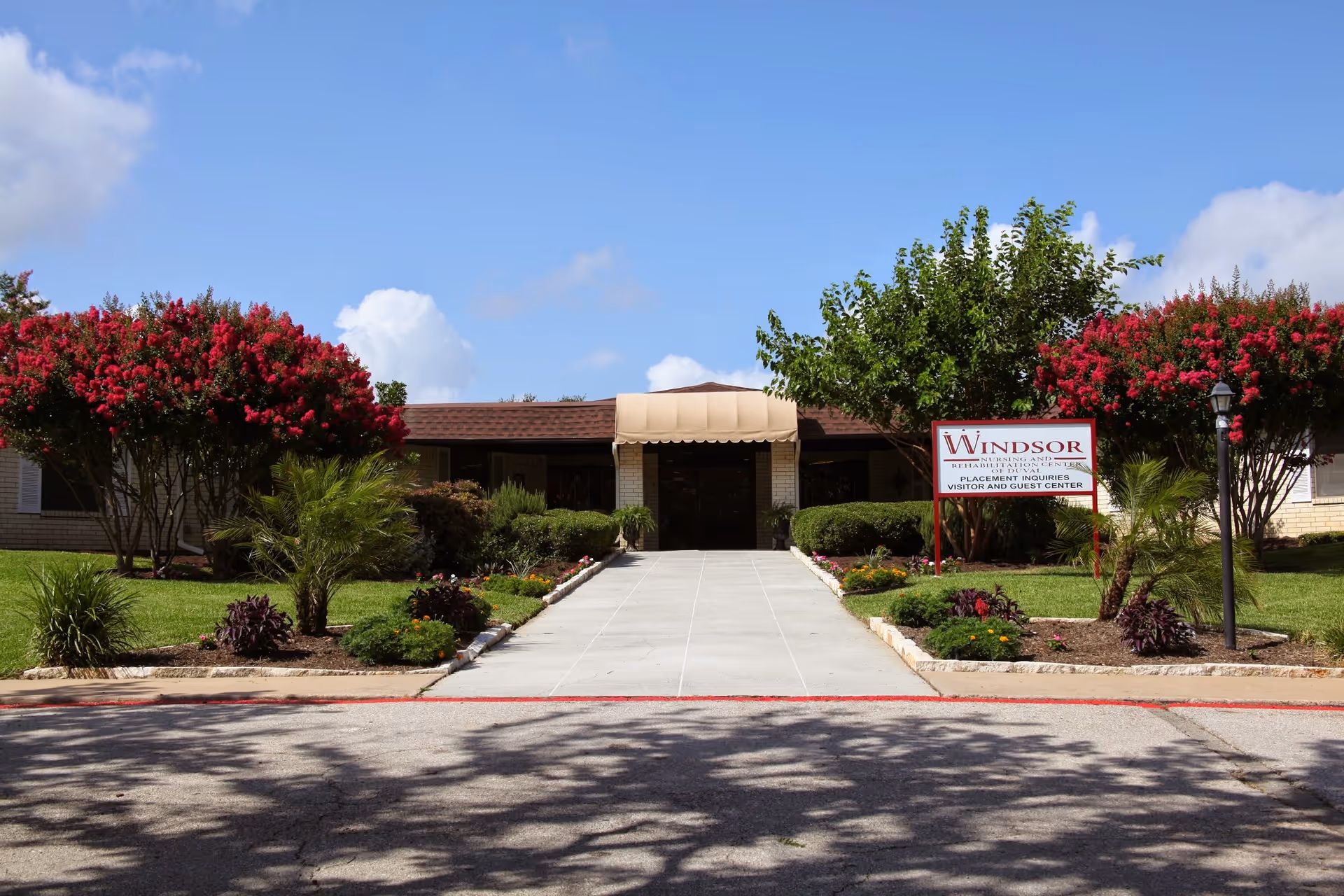 Front exterior view of Windsor Nursing and Rehabilitation Center of Duval with a paved walkway leading to the entrance, surrounded by well-maintained landscaping including green shrubs, small palm trees, and flowering trees with red blossoms under a blue sky with some clouds.