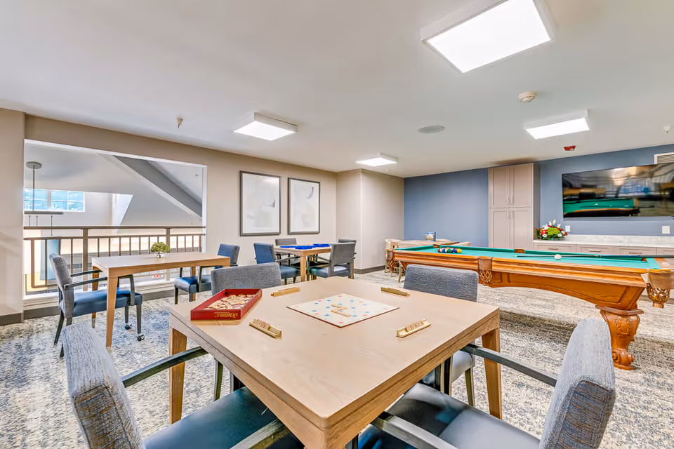 A bright and spacious game room in an assisted living facility featuring a pool table, multiple tables with chairs, and a Scrabble board game set up on the nearest table. The room has modern lighting, framed artwork on the walls, and a large TV mounted on a blue accent wall.