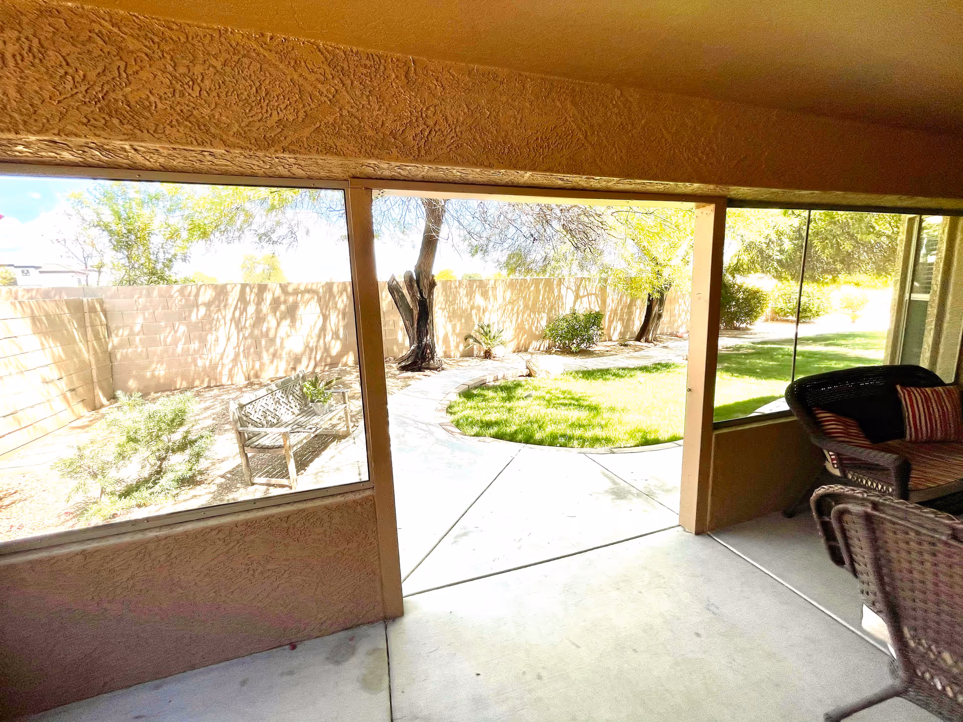 View from a covered patio area looking out onto a garden with a curved concrete pathway, green grass, trees, and a wooden bench along a beige brick wall. The patio has wicker chairs with striped cushions.