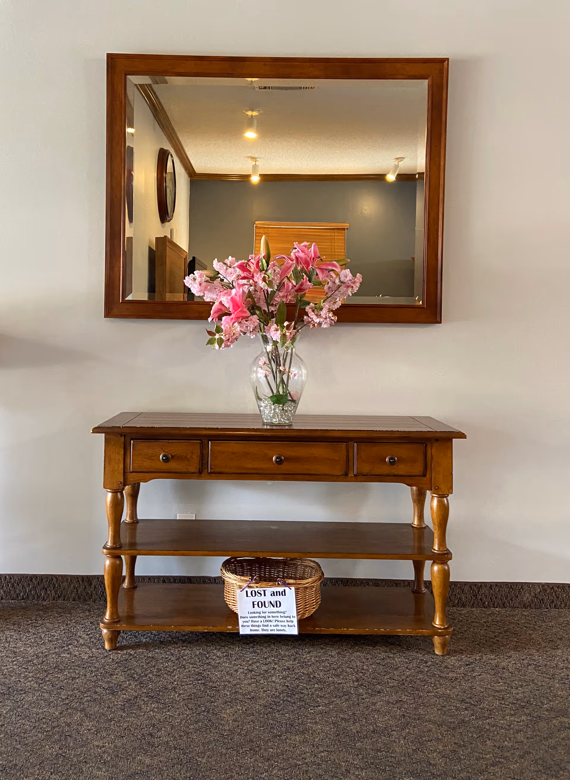 A wooden console table with three drawers and two shelves stands against a beige wall. On top of the table is a clear glass vase filled with pink flowers. Above the table hangs a large wooden-framed mirror reflecting ceiling lights and part of the room. On the bottom shelf of the table is a wicker basket with a sign labeled 'LOST and FOUND'.