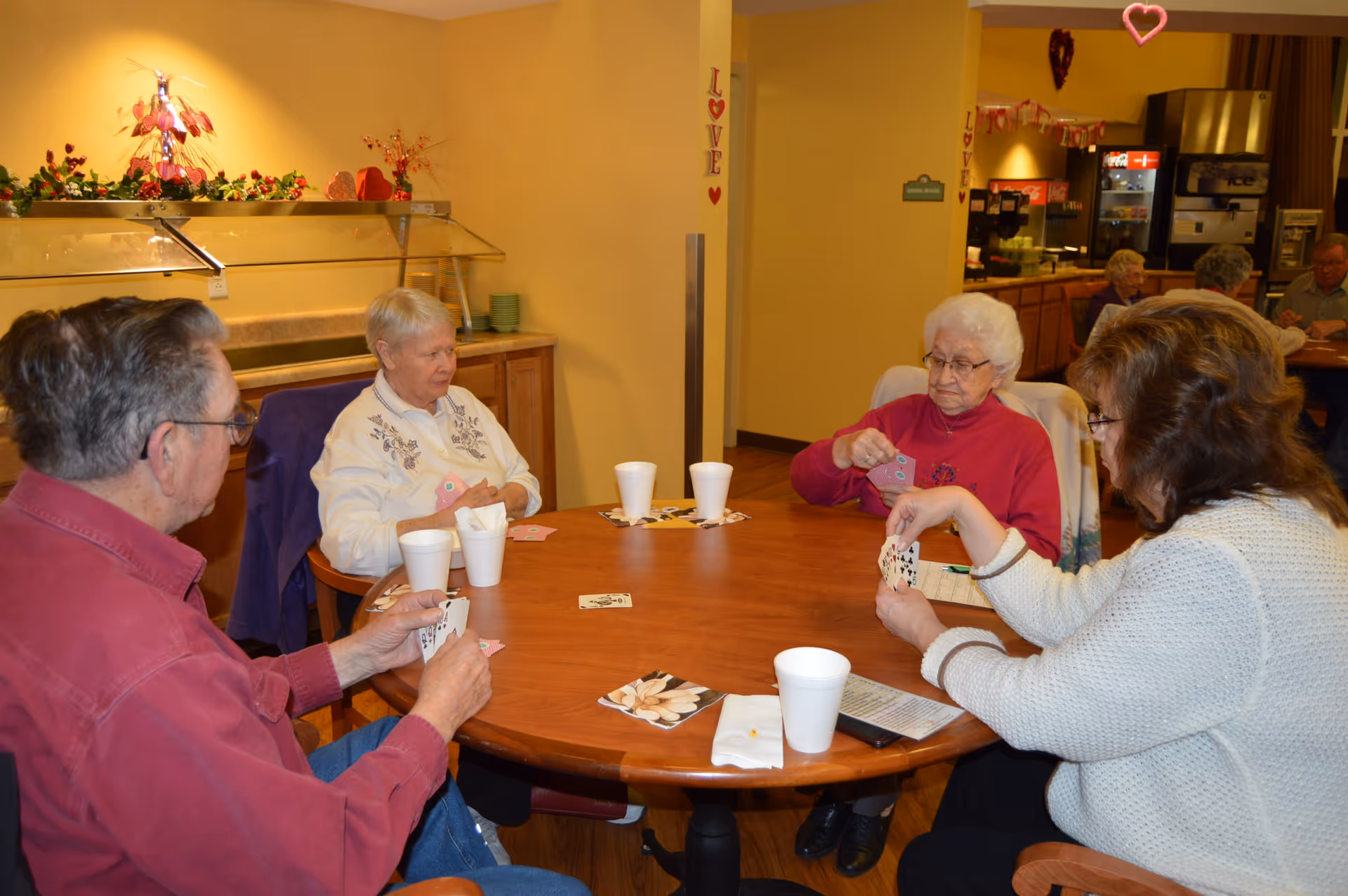 Four elderly people sitting around a round wooden table playing cards in a well-lit room decorated with Valentine-themed decorations. There are white disposable cups and napkins on the table. In the background, more elderly people are seated at tables near a beverage station.