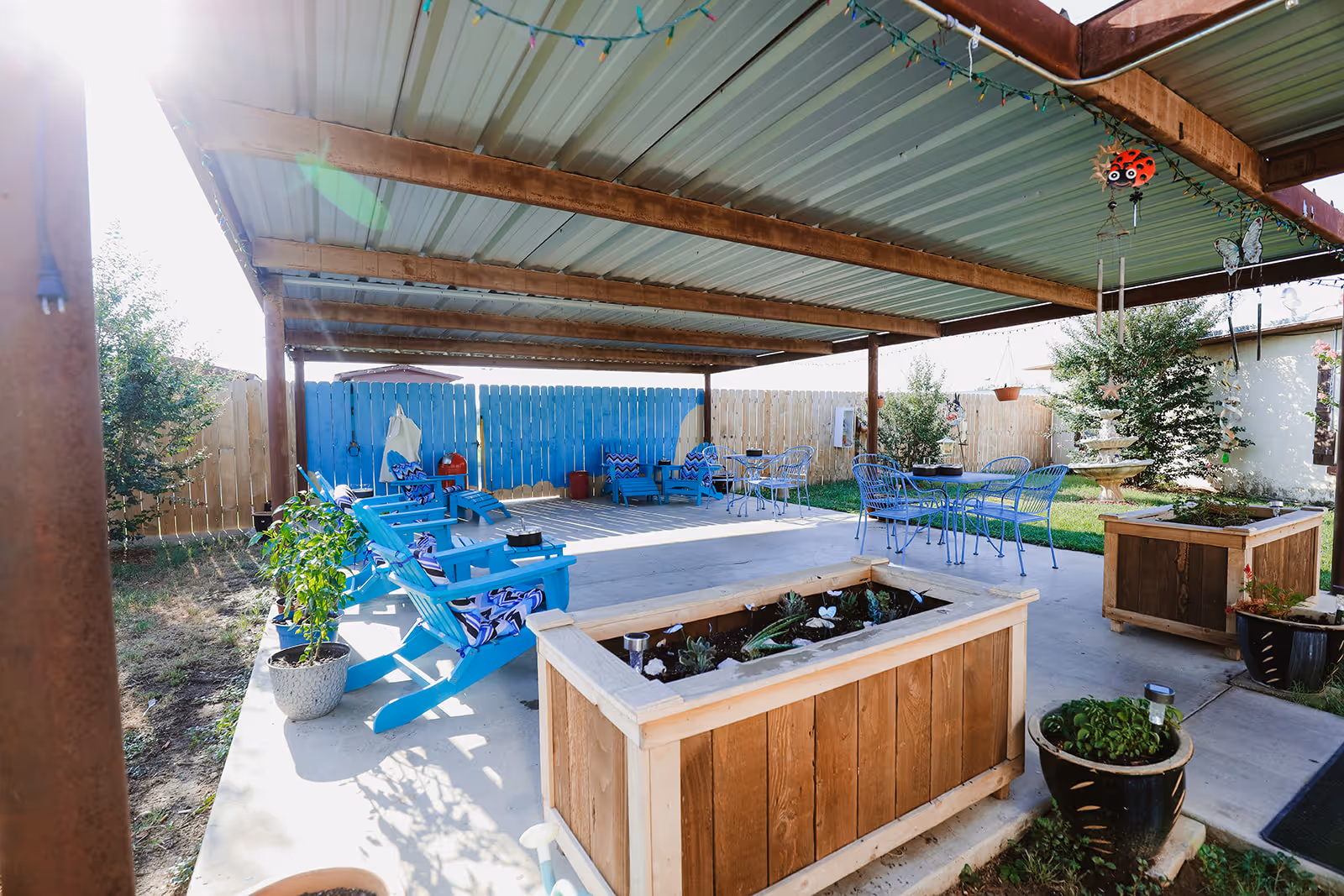 Covered outdoor patio area with blue Adirondack chairs, metal tables and chairs, potted plants, and wooden planters. The space is enclosed by a wooden fence and decorated with string lights and hanging ornaments.