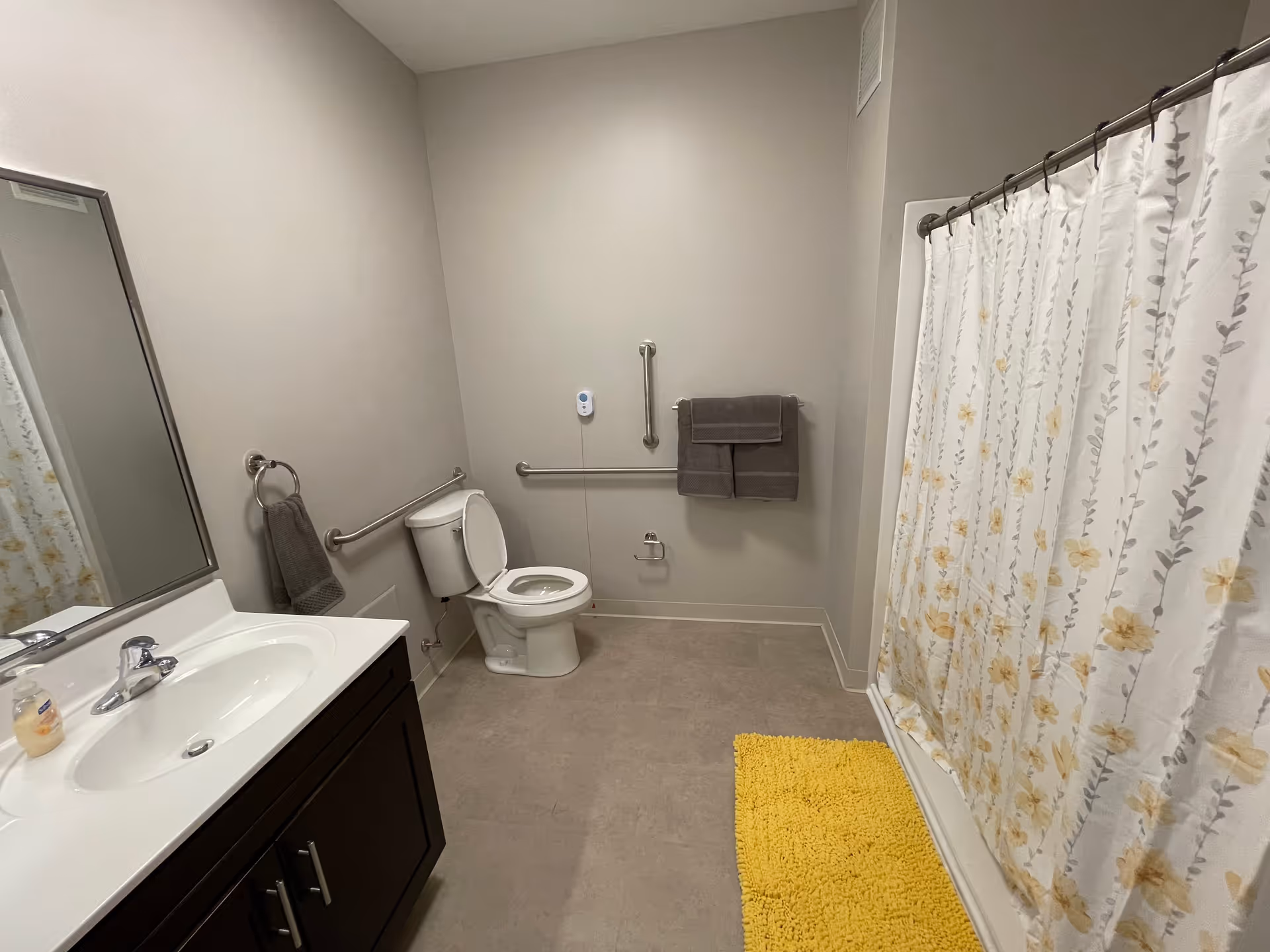 Bathroom featuring a sink and mirror, a toilet with grab bars, and a shower with a floral curtain and yellow bath mat.