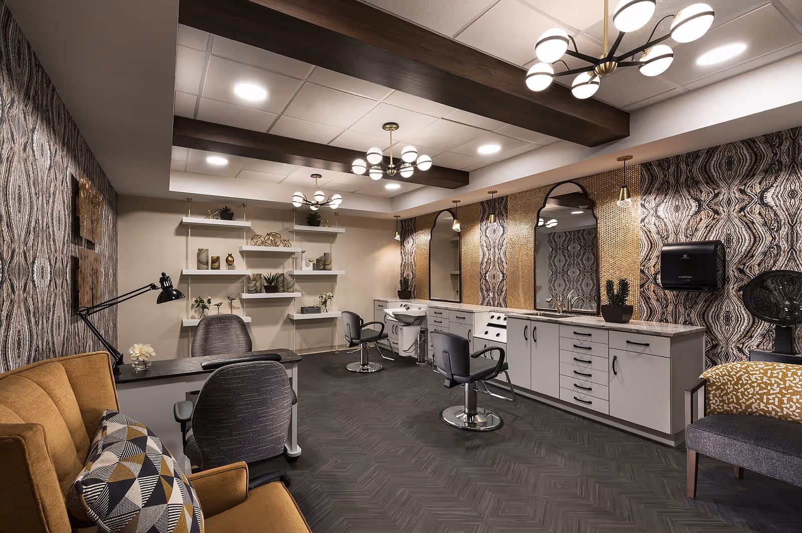 A stylish salon room with multiple chairs and mirrors along one wall. The room features patterned wallpaper, modern light fixtures, and a combination of shelves with decorative items and cabinetry with sinks. There is a mustard-colored armchair with a geometric patterned pillow and a gray chair with a yellow patterned cushion.