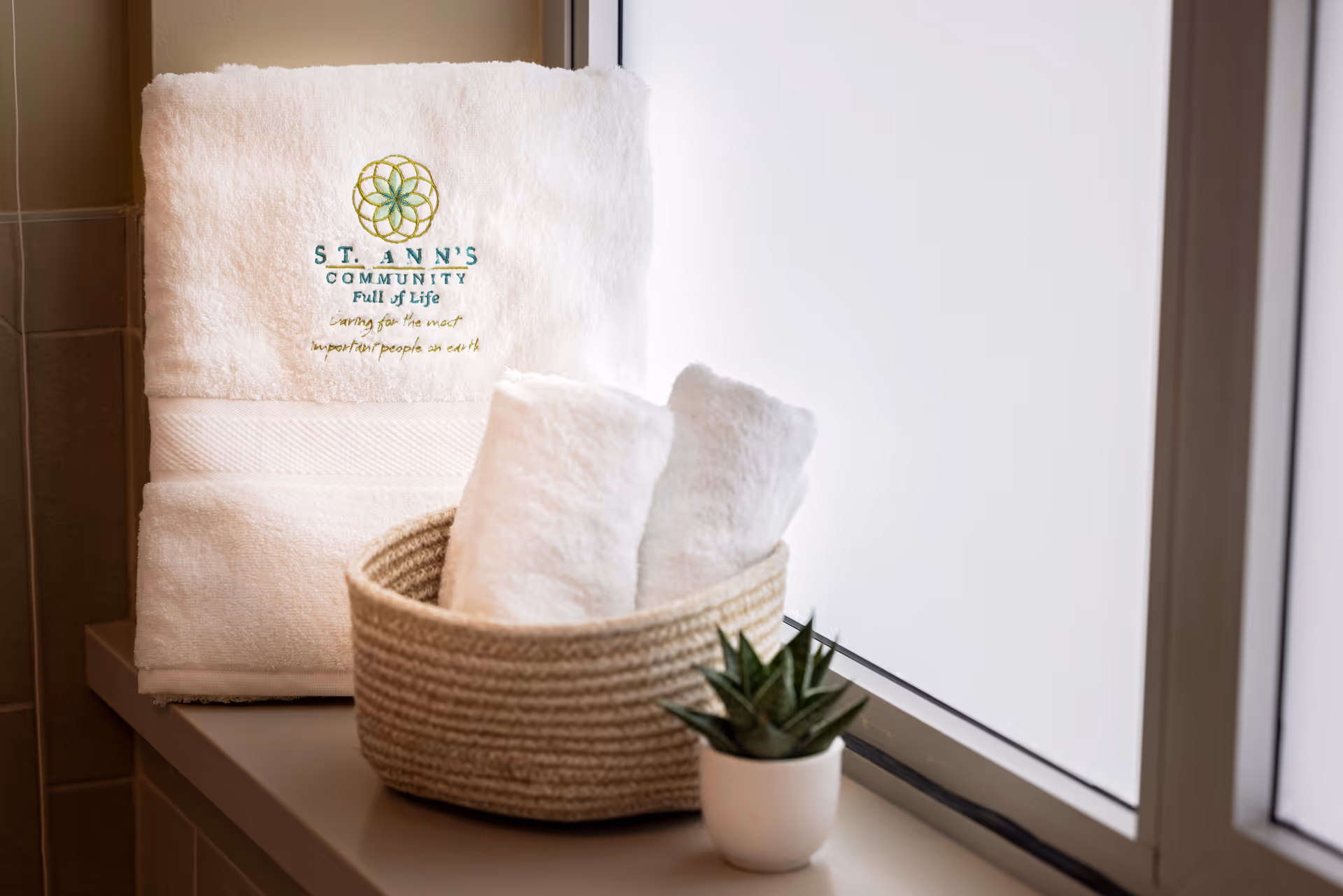 White embroidered "St. Ann's Community" towel on a windowsill with rolled towels in a woven basket and a small potted succulent.