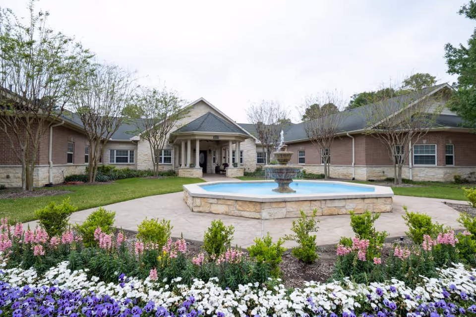 Front exterior of a single-story assisted living building with a central fountain, paved courtyard, and flowerbeds.