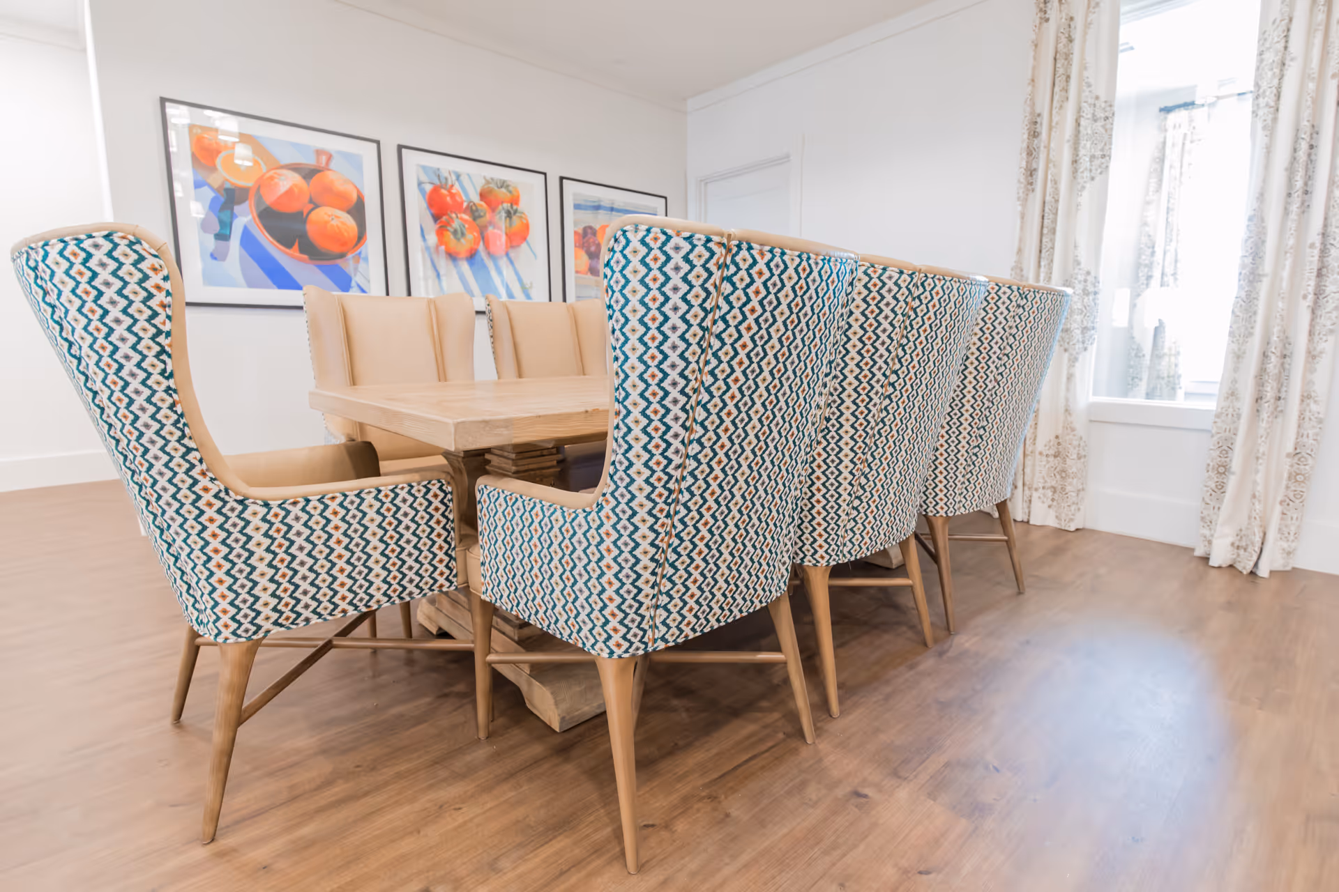 A dining room with a wooden table surrounded by eight upholstered chairs featuring a geometric pattern in blue, white, and tan. The room has light-colored walls, three framed paintings of fruit on the wall, wood flooring, and windows with patterned curtains allowing natural light to enter.