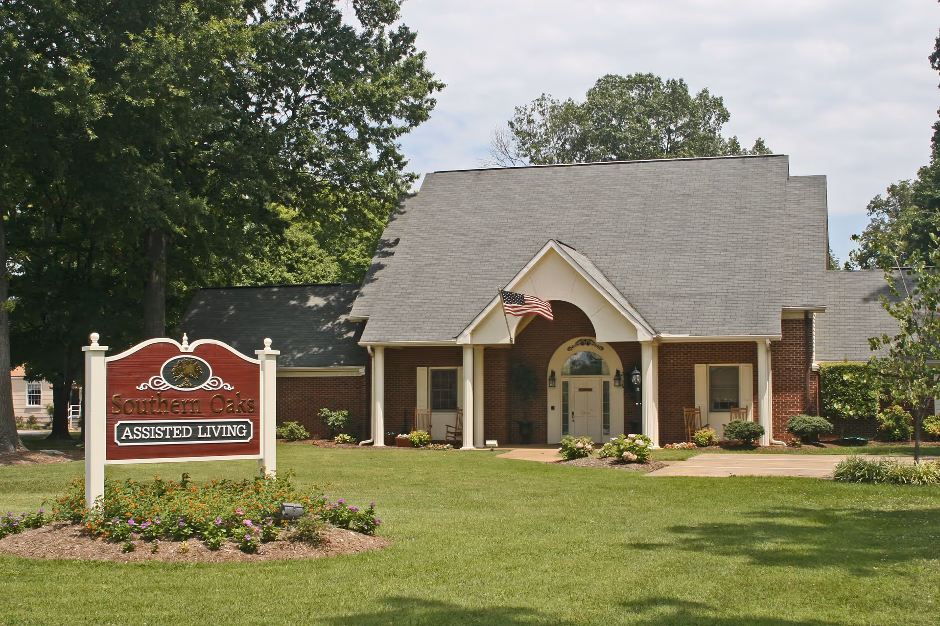 Front exterior view of Southern Oaks Assisted Living facility, featuring a brick building with a peaked roof, an American flag near the entrance, and a landscaped lawn with flowers and trees. A wooden sign in the foreground reads 'Southern Oaks Assisted Living'.