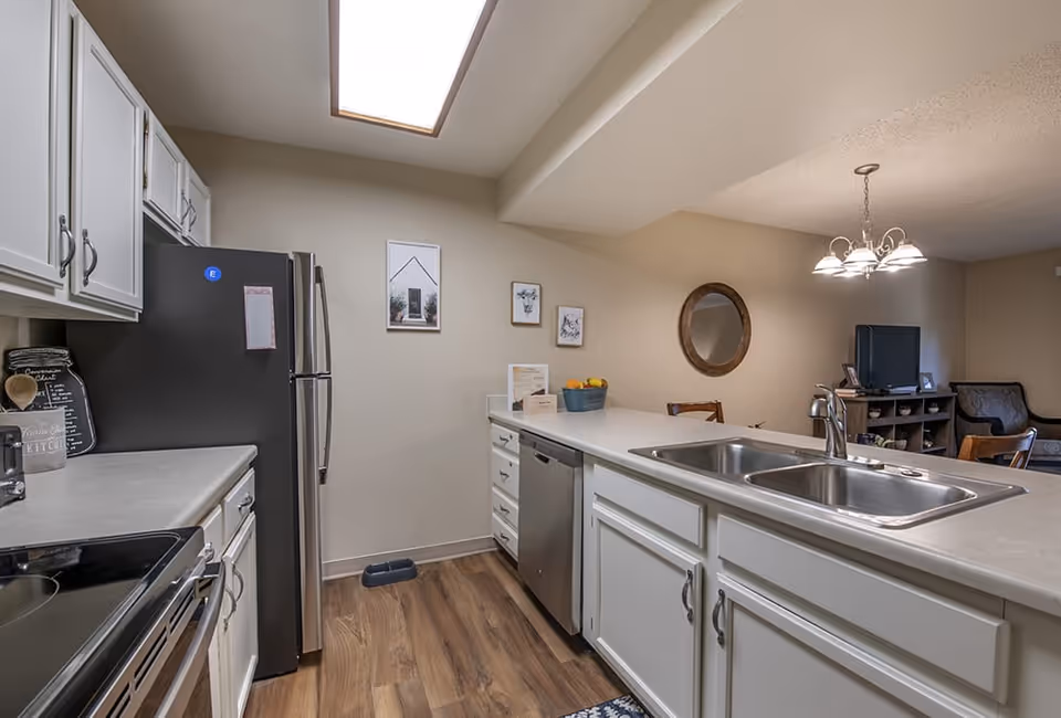 A modern kitchen with white cabinets, a double sink, stainless steel refrigerator and dishwasher, and wood flooring. The kitchen opens into a living area with a round mirror on the wall, a chandelier, and a TV on a stand.