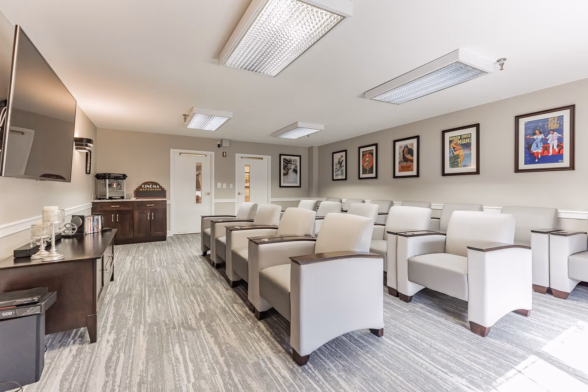 A small theater room with two rows of beige armchairs facing a large flat-screen TV mounted on the wall. The room has light gray walls adorned with framed vintage movie posters. There is a dark wooden cabinet with a popcorn machine and a sign that reads 'Cinema Always Showing' near two white doors at the back.