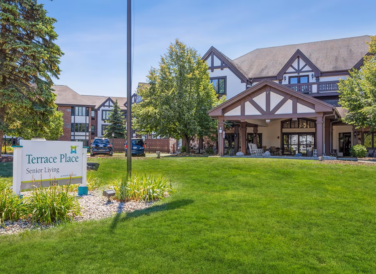 Exterior view of a senior living facility with a large green lawn, trees, and a building featuring Tudor-style architecture. A sign in the foreground reads 'Terrace Place Senior Living.' Several cars are parked near the building under a clear blue sky.
