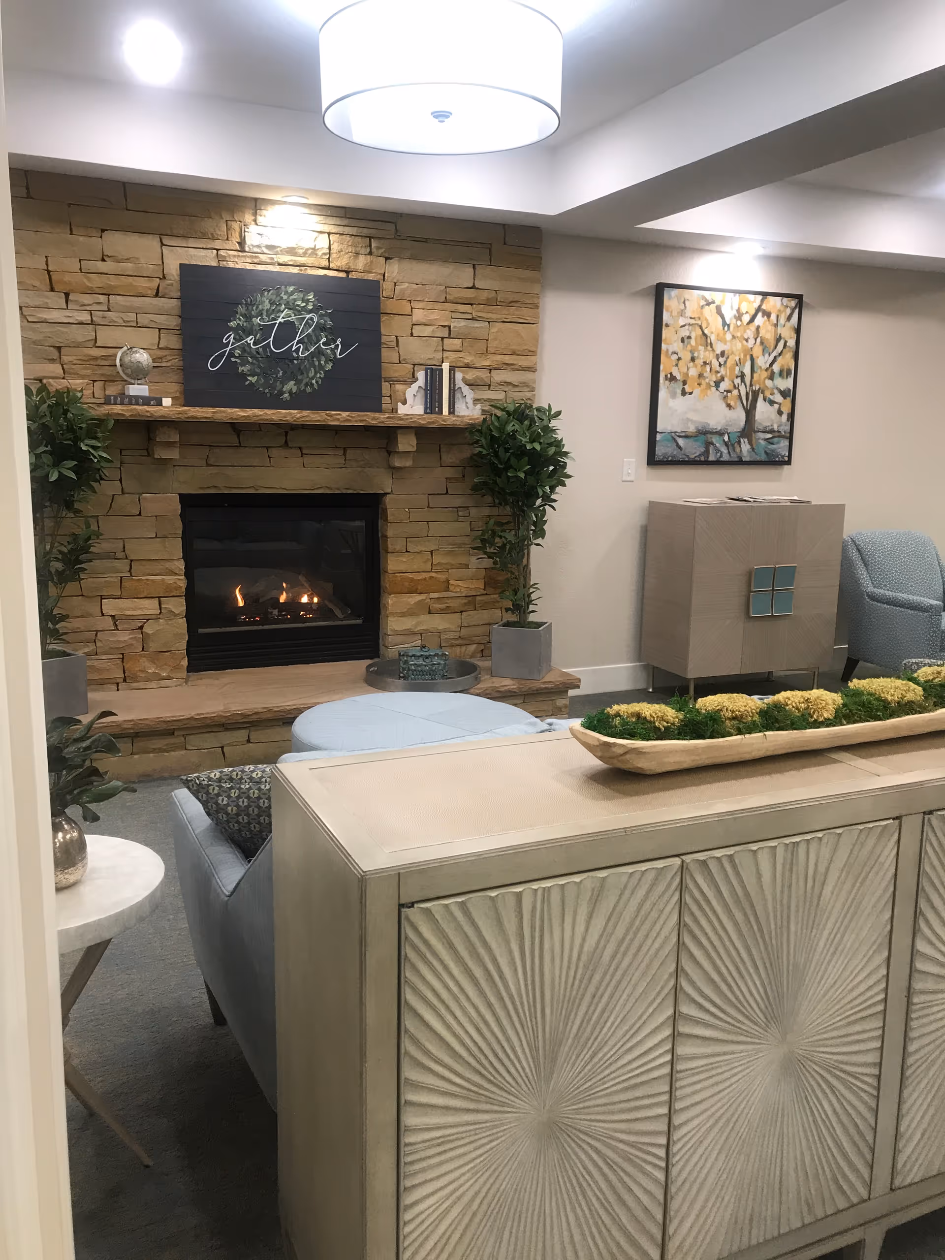 A cozy living room area with a stone fireplace that has a fire burning. Above the fireplace is a wooden shelf with decorative items and a sign that says 'gather' with a wreath design. There are two potted plants on either side of the fireplace. In the foreground, there is a light-colored cabinet with a decorative tray holding green and yellow moss balls. To the right, there is a light blue armchair and a small cabinet with a colorful abstract painting hanging above it. The room is softly lit with a ceiling light fixture.