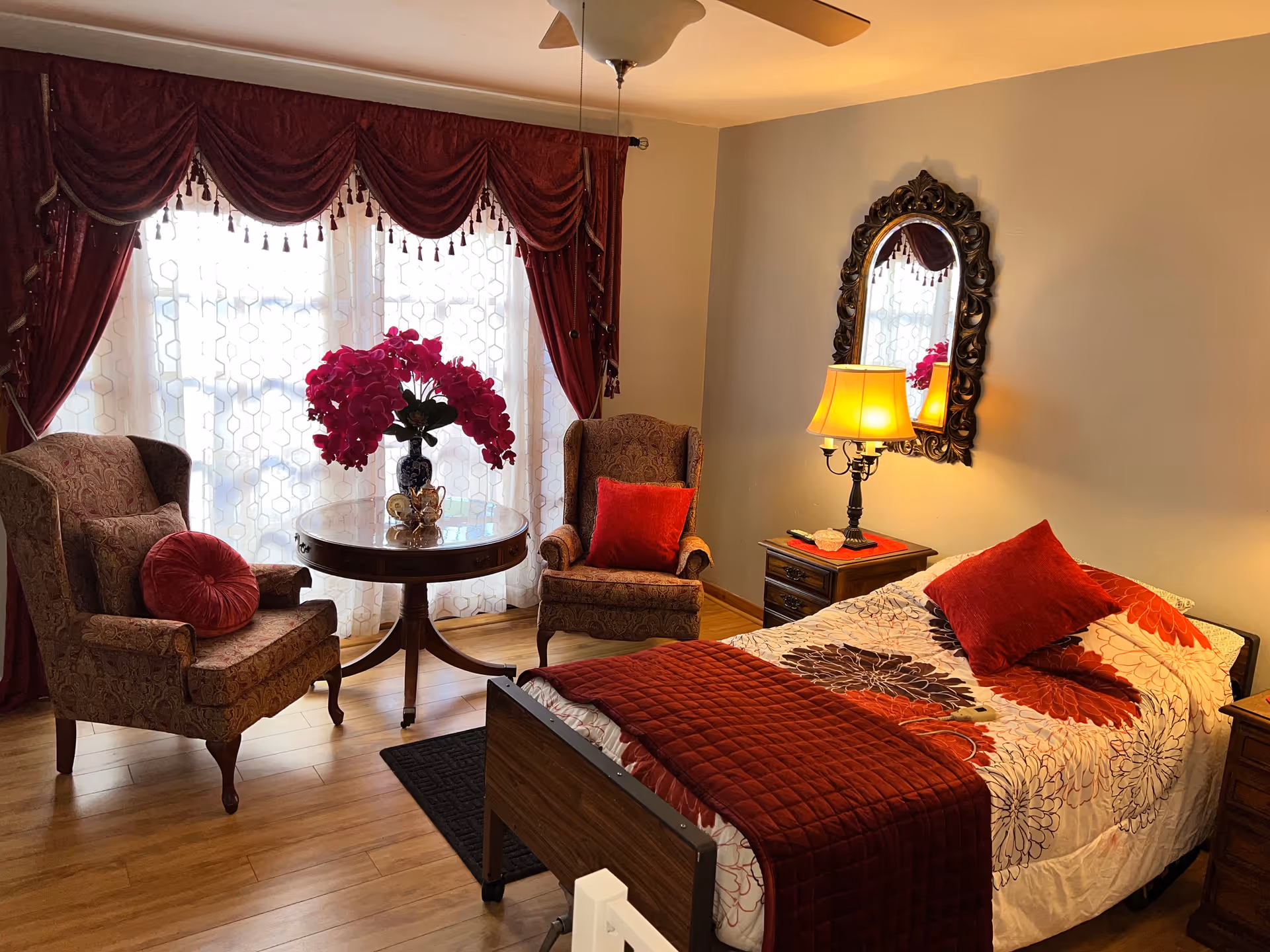 A cozy bedroom with a single bed covered in a floral bedspread and red pillows. Next to the bed is a wooden nightstand with a lamp and a decorative mirror hanging on the wall above it. Two upholstered armchairs with red cushions flank a round wooden table holding a vase with bright pink flowers. The room has wooden flooring and large windows with sheer white curtains and red drapes.