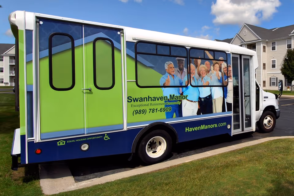 A green and blue Swanhaven Manor shuttle bus with a photo of smiling seniors on its side parked outside a retirement community building.