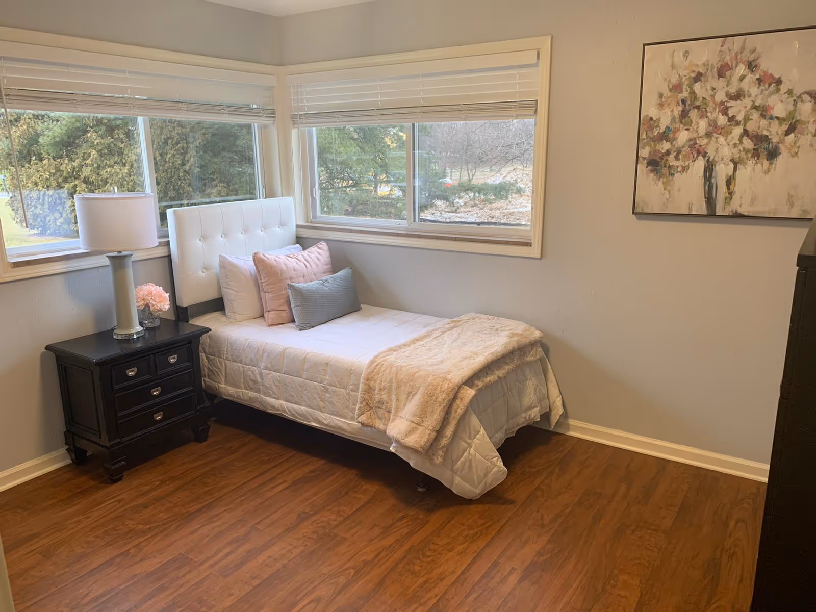 A small bedroom with a single bed against the corner windows. The bed has white bedding, a beige throw blanket, and three pillows in white, pink, and blue. Next to the bed is a black nightstand with three drawers, a white lamp, and a small pink flower arrangement. The room has wooden flooring, light-colored walls, and a large floral painting on the wall.